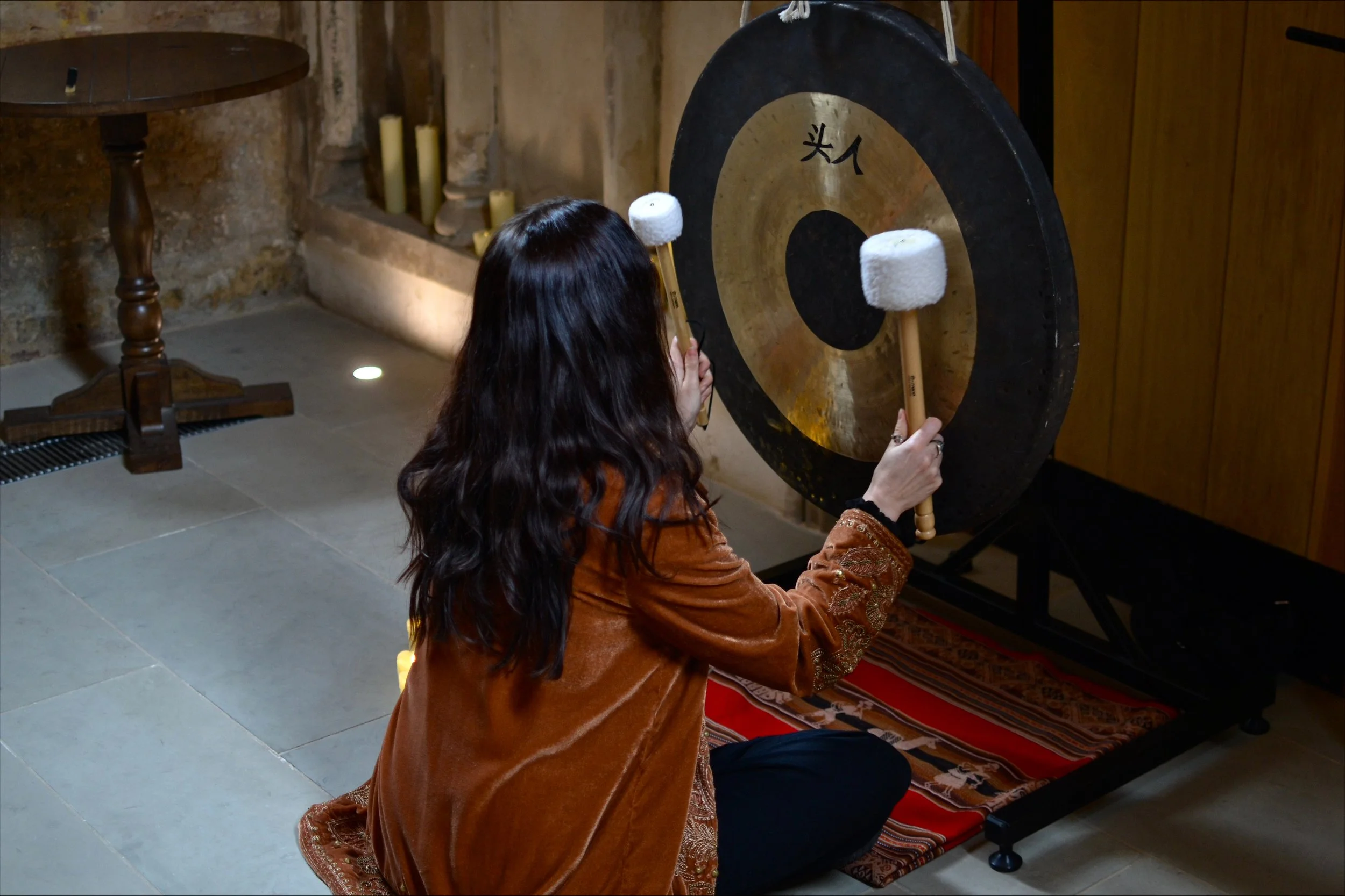 A woman with long, dark, wavy hair, wearing an orange-brown velvet jacket, strikes a large gong with two mallets in a rustic room with stone and wood walls, candles, and a small table.