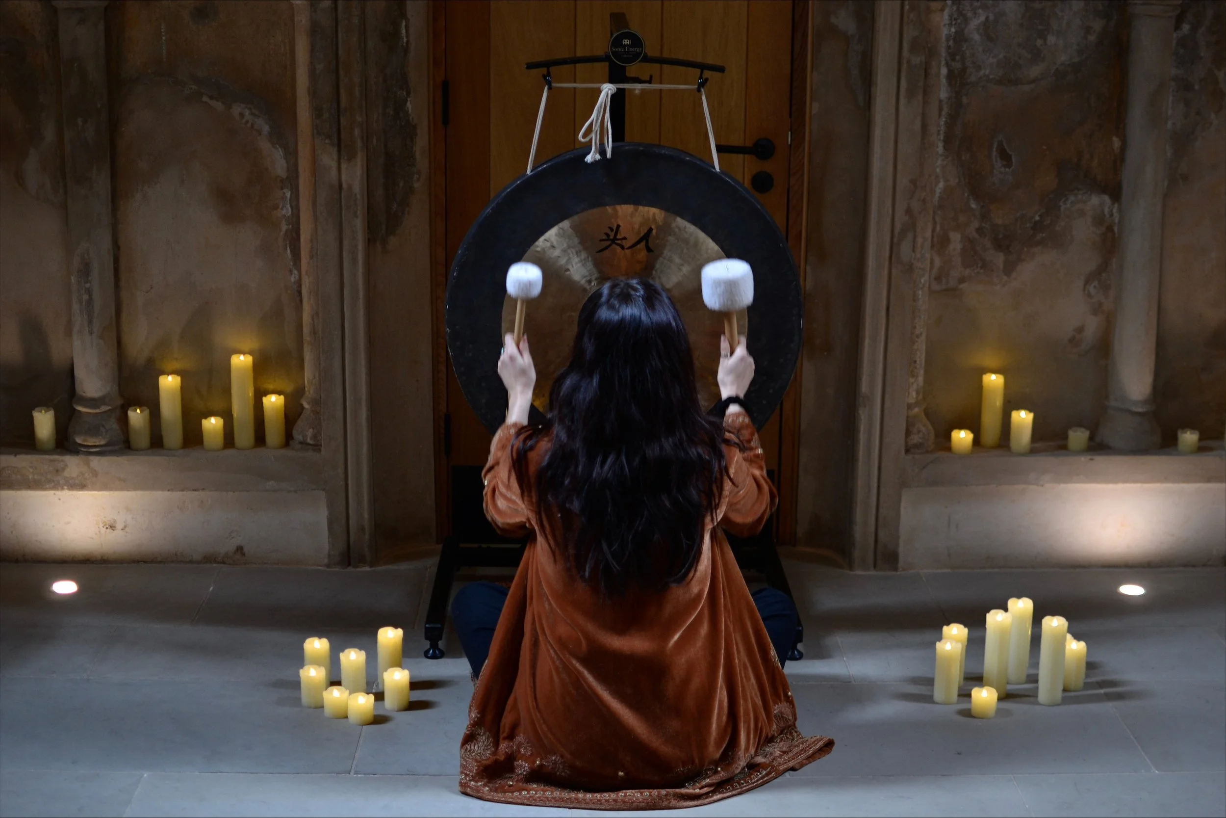 A woman with long black hair, wearing a brown velvet jacket, sitting cross-legged in front of a large gong, holding two mallets with white heads, in a room with lit candles.