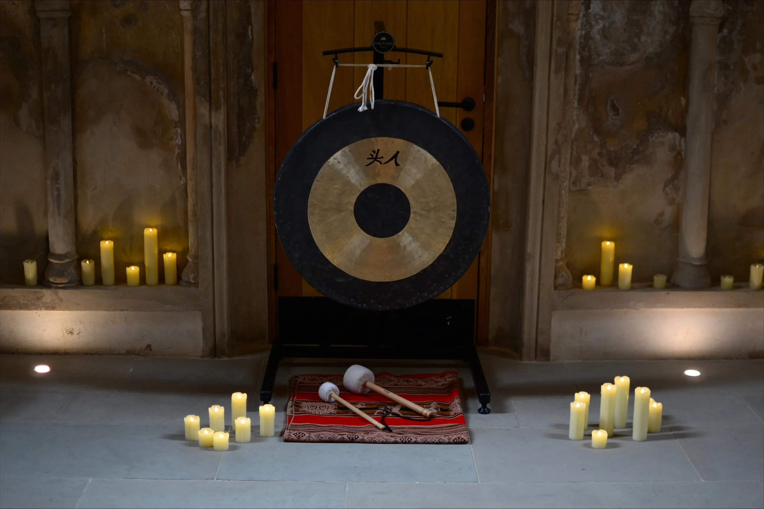 A large gong with traditional Chinese characters, surrounded by lit candles and percussion mallets placed on a patterned cloth, in a dimly lit room with stone walls.