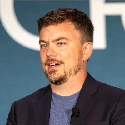 A man with short brown hair and a beard speaking at an event, wearing a dark blazer and a blue shirt, with a microphone clipped to his shirt.