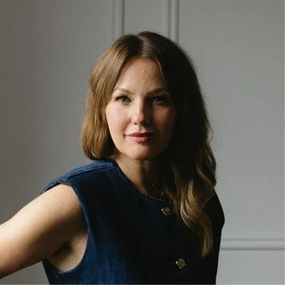 A woman with shoulder-length brown hair wearing a sleeveless denim jacket, standing indoors with white paneled walls.