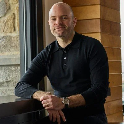 Man with a shaved head and beard sitting by a window in a wooden-paneled room, wearing a black long-sleeve shirt.