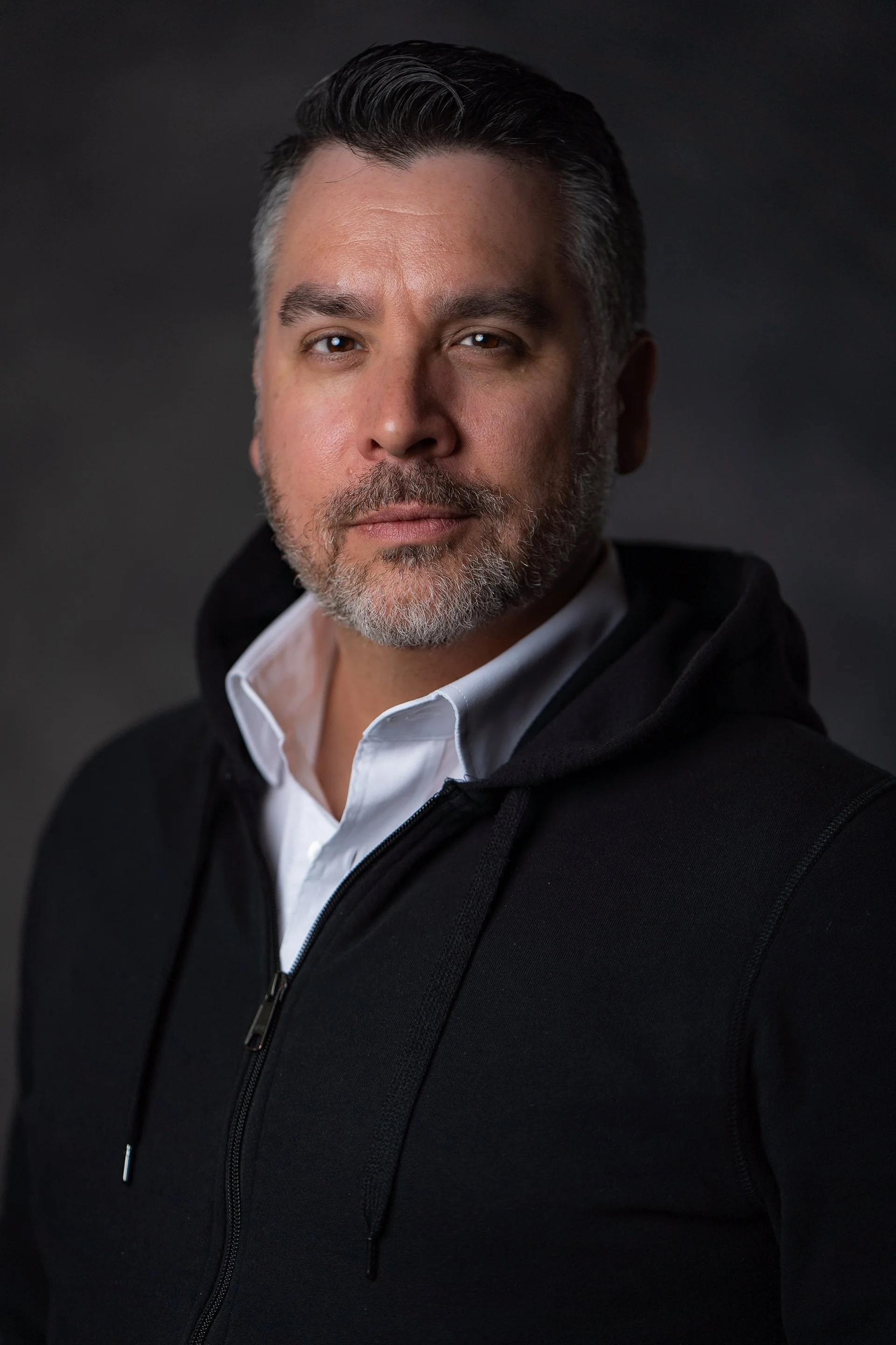 Portrait of a middle-aged man with short dark hair and a beard, wearing a white collared shirt and a black zip-up hoodie, against a dark background.