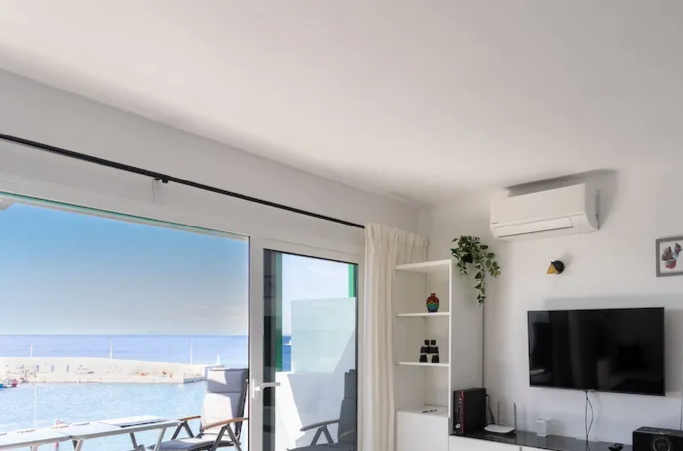 Living room with large sliding glass door overlooking a beach and sea, white walls, flat-screen TV, air conditioner, white bookshelf with decorative items, potted plant, and a balcony with outdoor chairs.