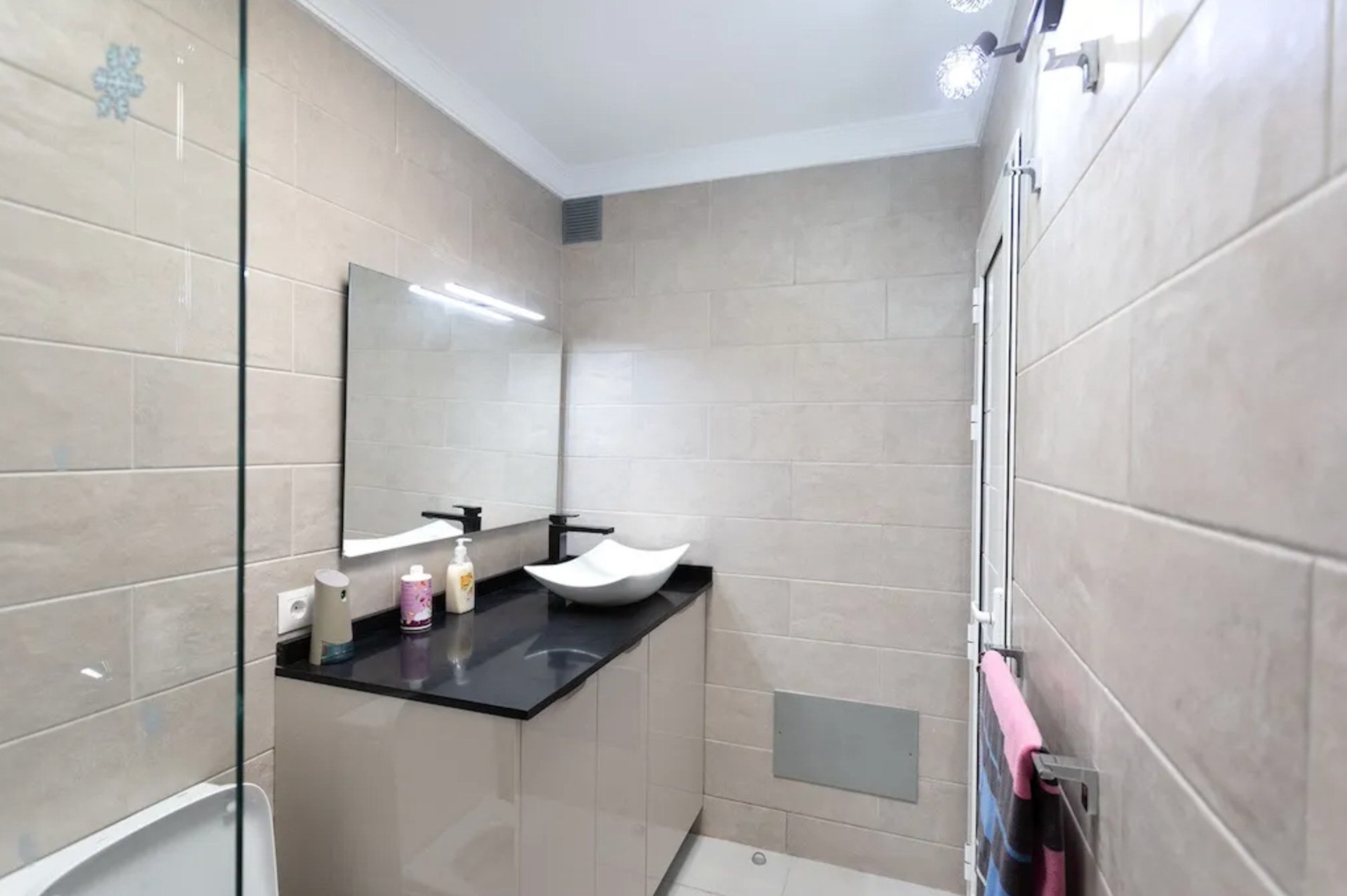 Modern bathroom with beige tiled walls, a black countertop with a white vessel sink, a large mirror, and a towel rack with pink towels. There are various toiletries on the counter, and a glass shower door is partially visible on the left.