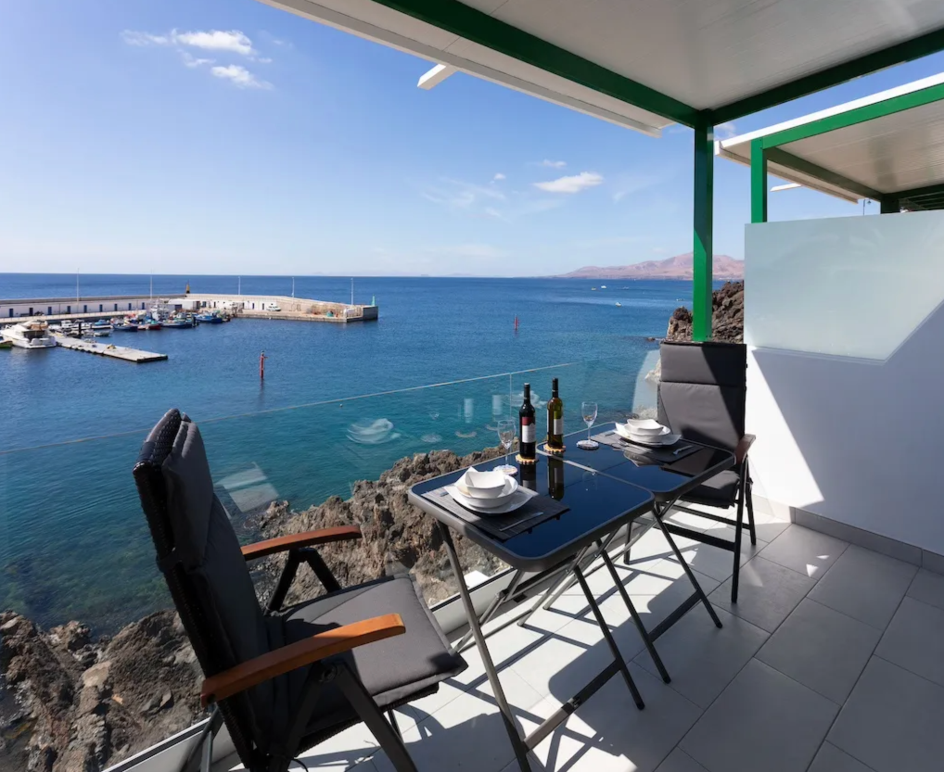 A balcony overlooking a marina with boats, with a dining table set with plates, wine bottles, and glasses, overlooking blue ocean and distant land on a sunny day.