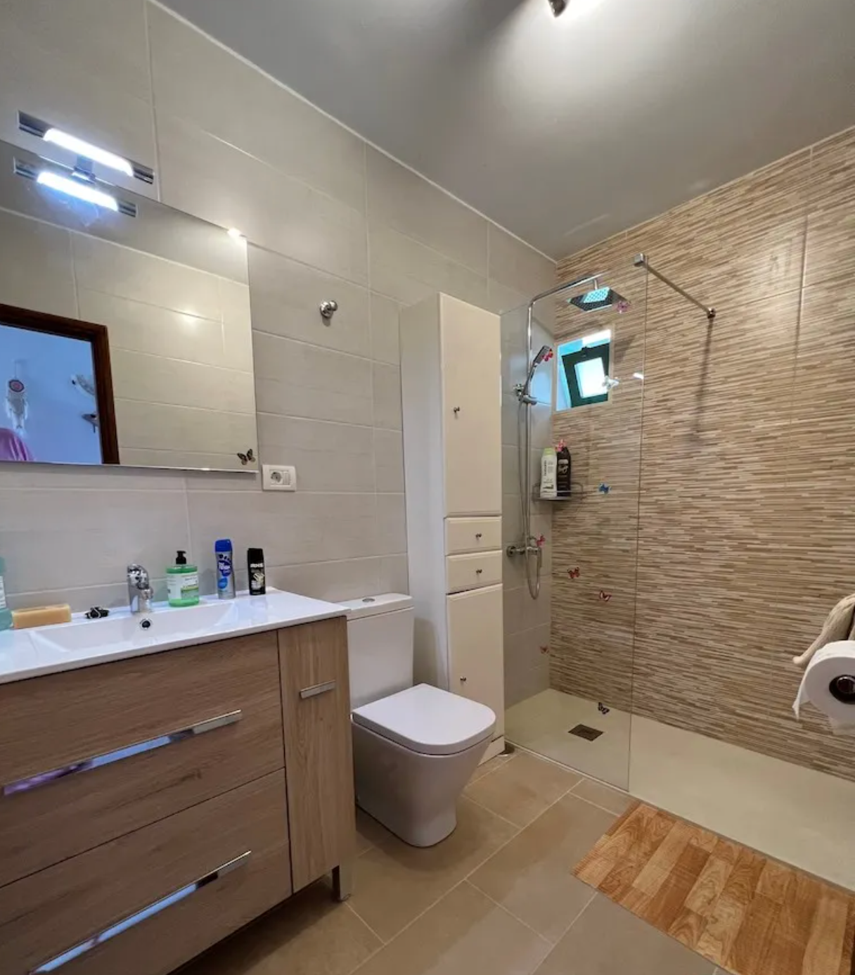 Modern bathroom with wooden vanity, white sink, toilet, wall mirror, glass shower area with beige striped tiles, small window, and a bath mat.