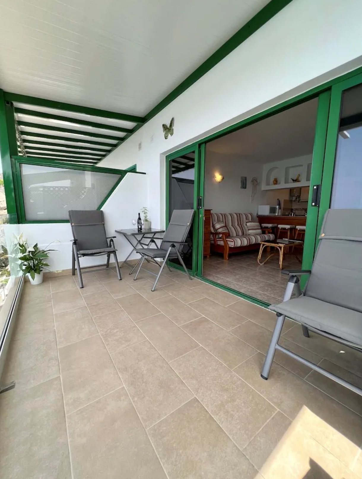 Balcony with two gray chairs, a small table with a bottle and glass, potted plants, and a sliding glass door leading inside a living room with a striped sofa and wooden furniture.