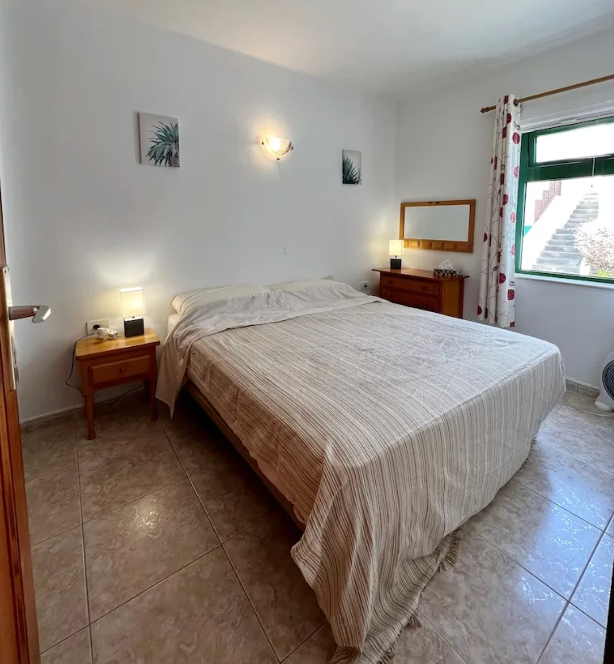 A bedroom with a large bed covered in a striped beige and brown bedspread, two small wooden nightstands with lamps, a wooden dresser with a mirror, a window with patterned curtains, tiled floor, and some wall art.