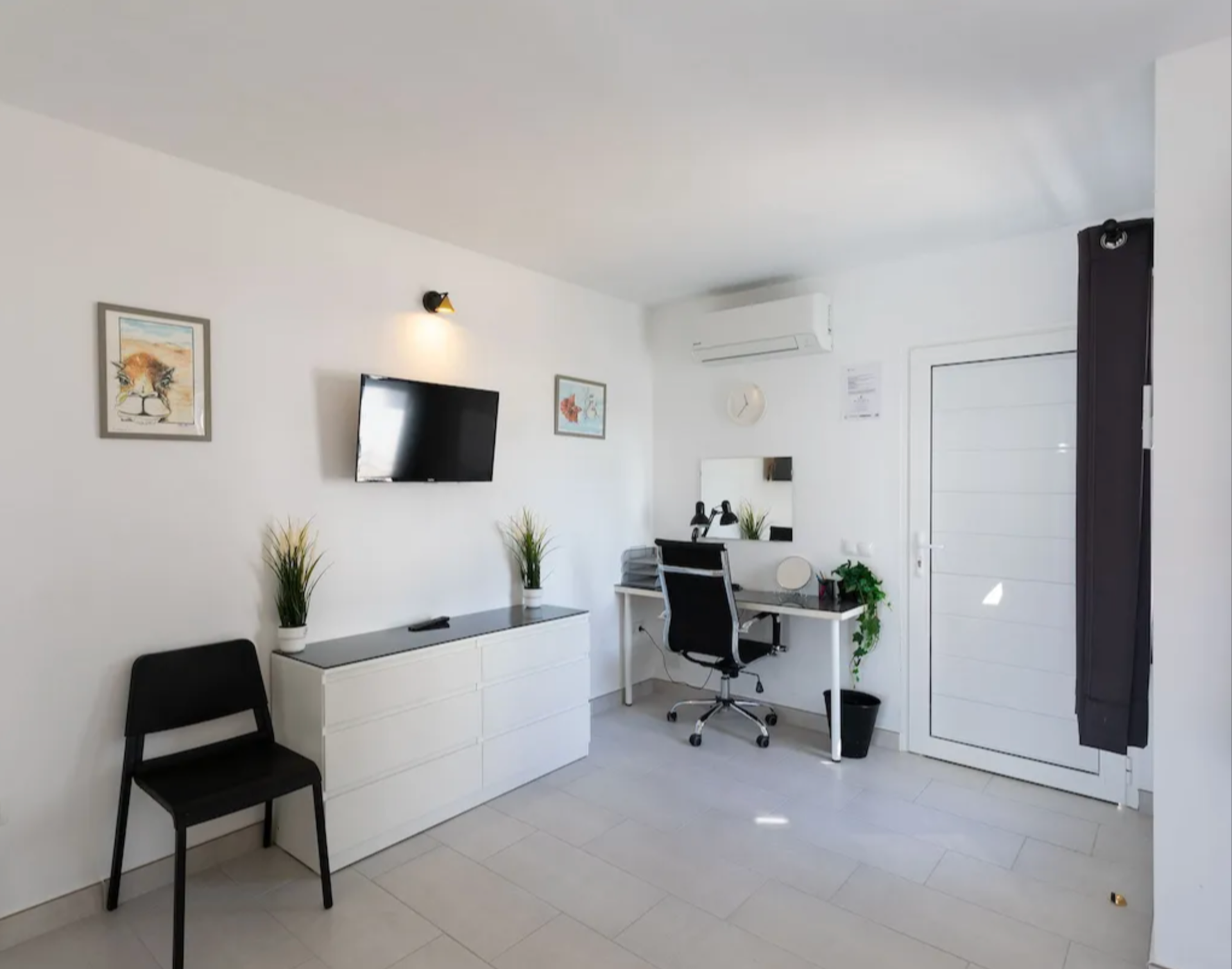 A modern, minimalist room with a white wall, featuring a wall-mounted TV, two framed animal artworks, a white sideboard with potted plants, a black chair, a desk with a black office chair, a mirror, a small potted plant, and an air conditioning unit.