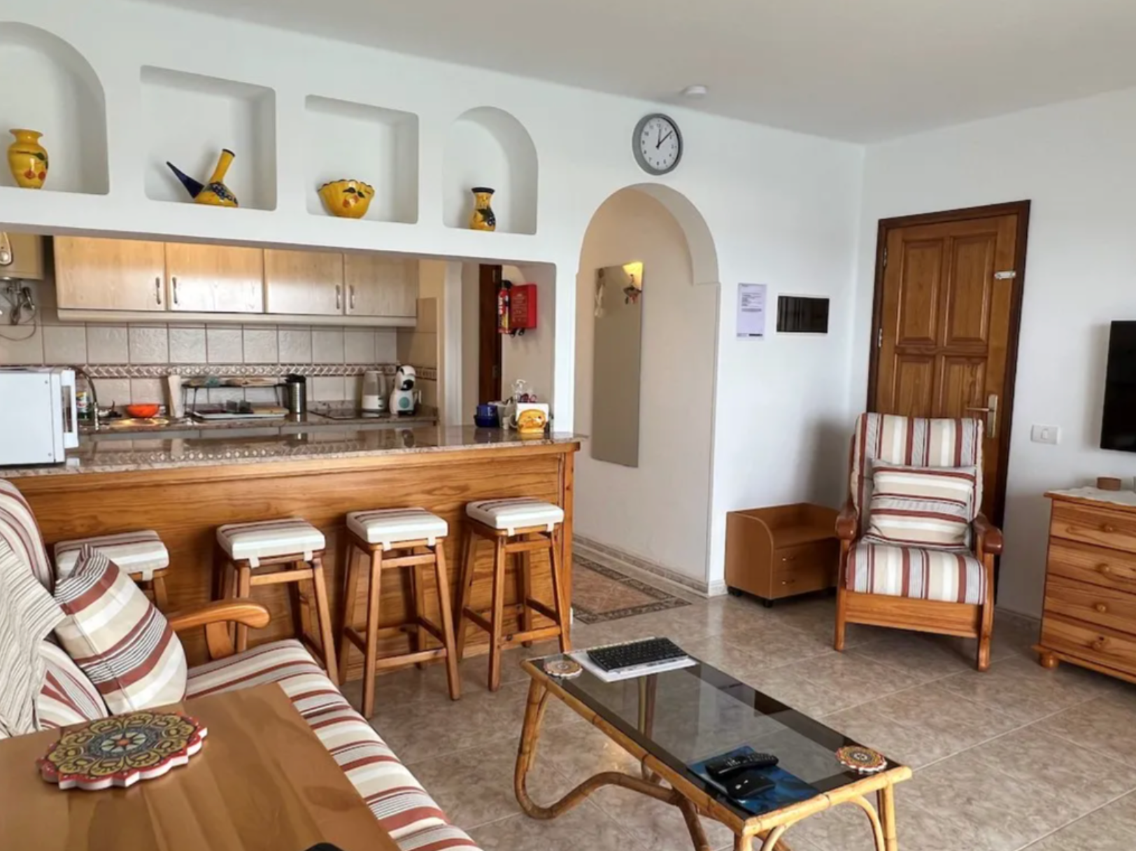 Living room and kitchen with wooden furniture, striped upholstery, island with stools, and decorative yellow vases on shelves.
