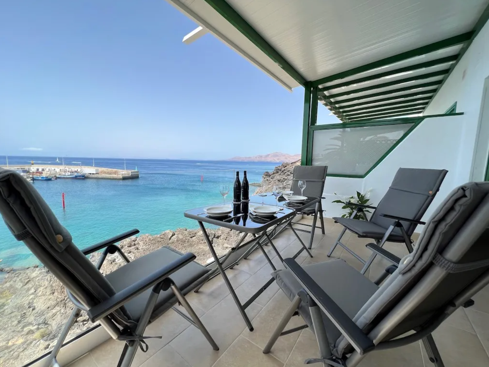 A balcony with a sea view, set with a small table, four chairs, bottles of wine, wine glasses, plates, and a plant.