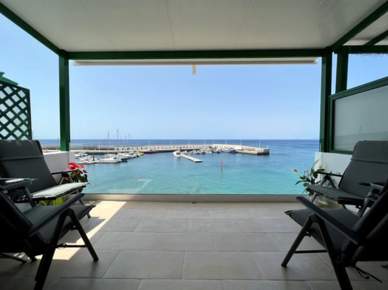 Balcony with chairs overlooking a marina with sailboats and the ocean, under a roof with green accents.