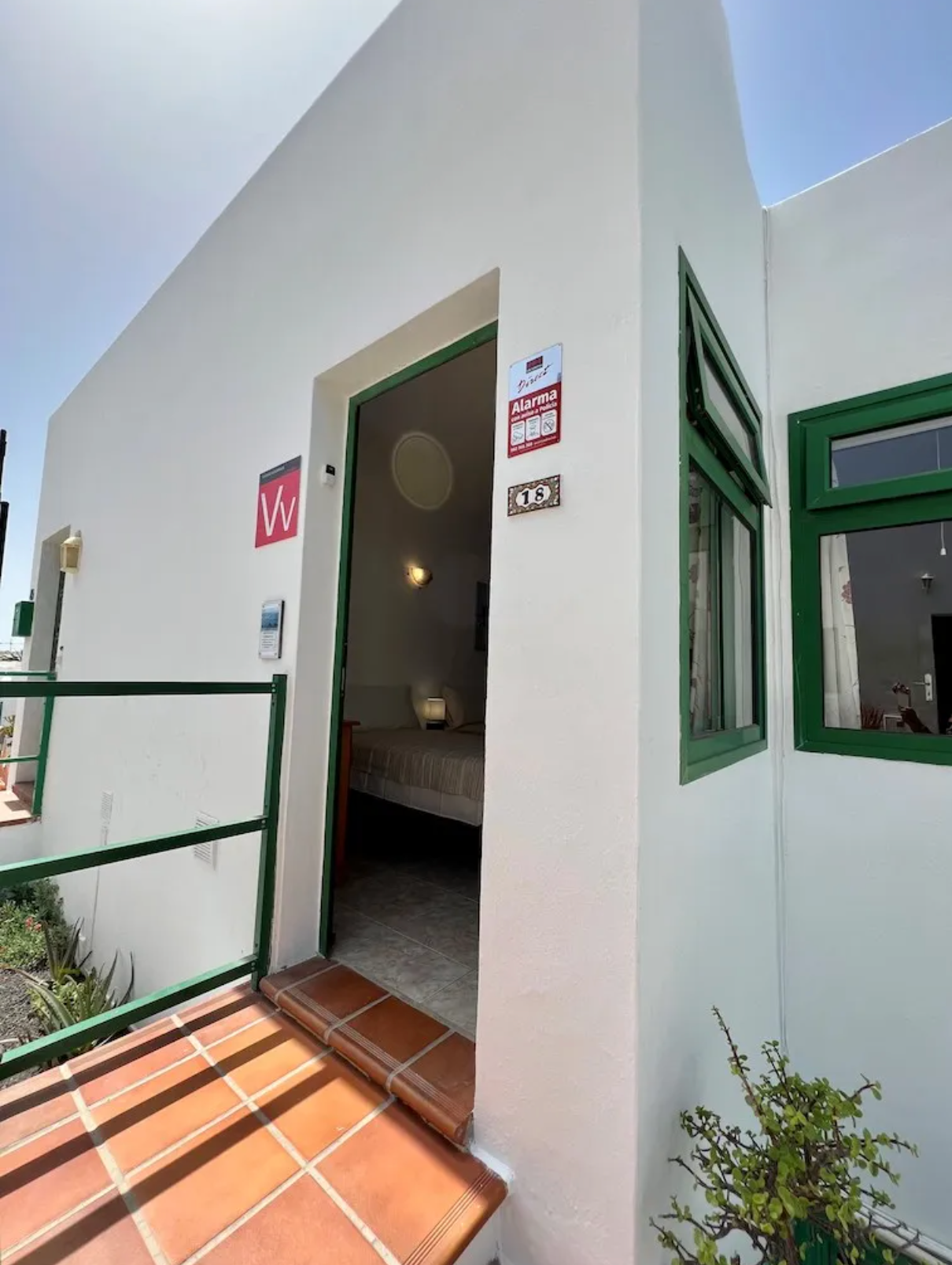 Exterior of a white building with green window frames, an open doorway, and a small tiled porch, with plants and a clear blue sky.