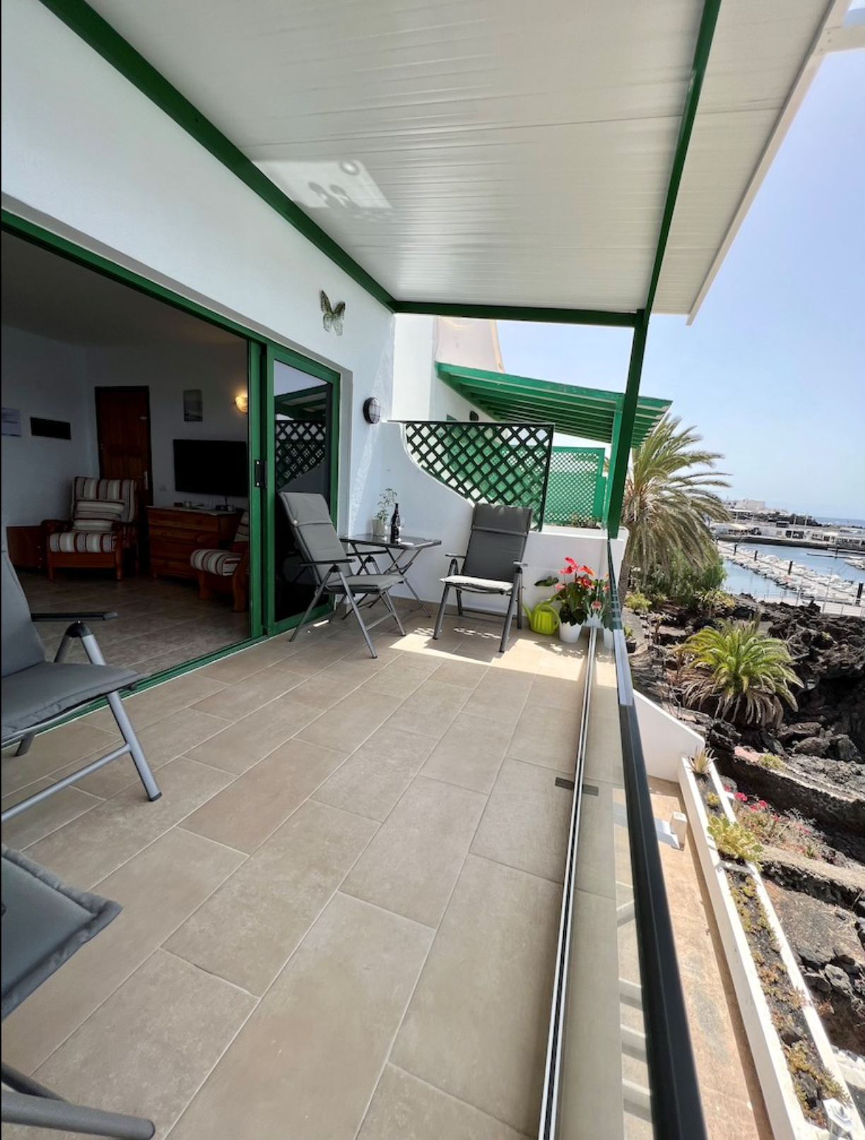 Balcony with chairs, potted plants, and a view of a marina with boats and palm trees.