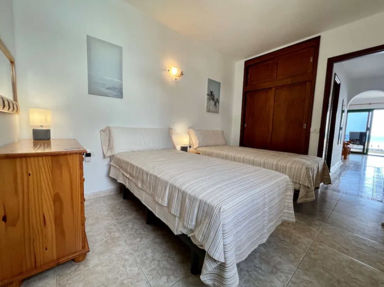 Room with two twin beds covered in beige striped bedspreads, wooden nightstand with lamp between the beds, and tropical artwork on the white wall. There is a wooden closet and a doorway leading to a balcony or patio area with outdoor furniture and a 