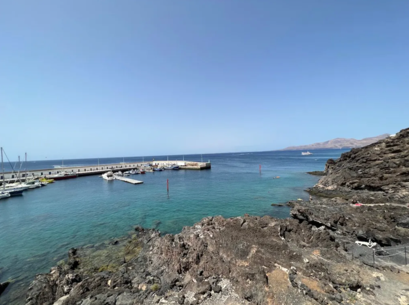 A marina with boats docked along a concrete pier, surrounded by turquoise water and rugged rocky coastline, under clear blue skies with distant mountains on the horizon.
