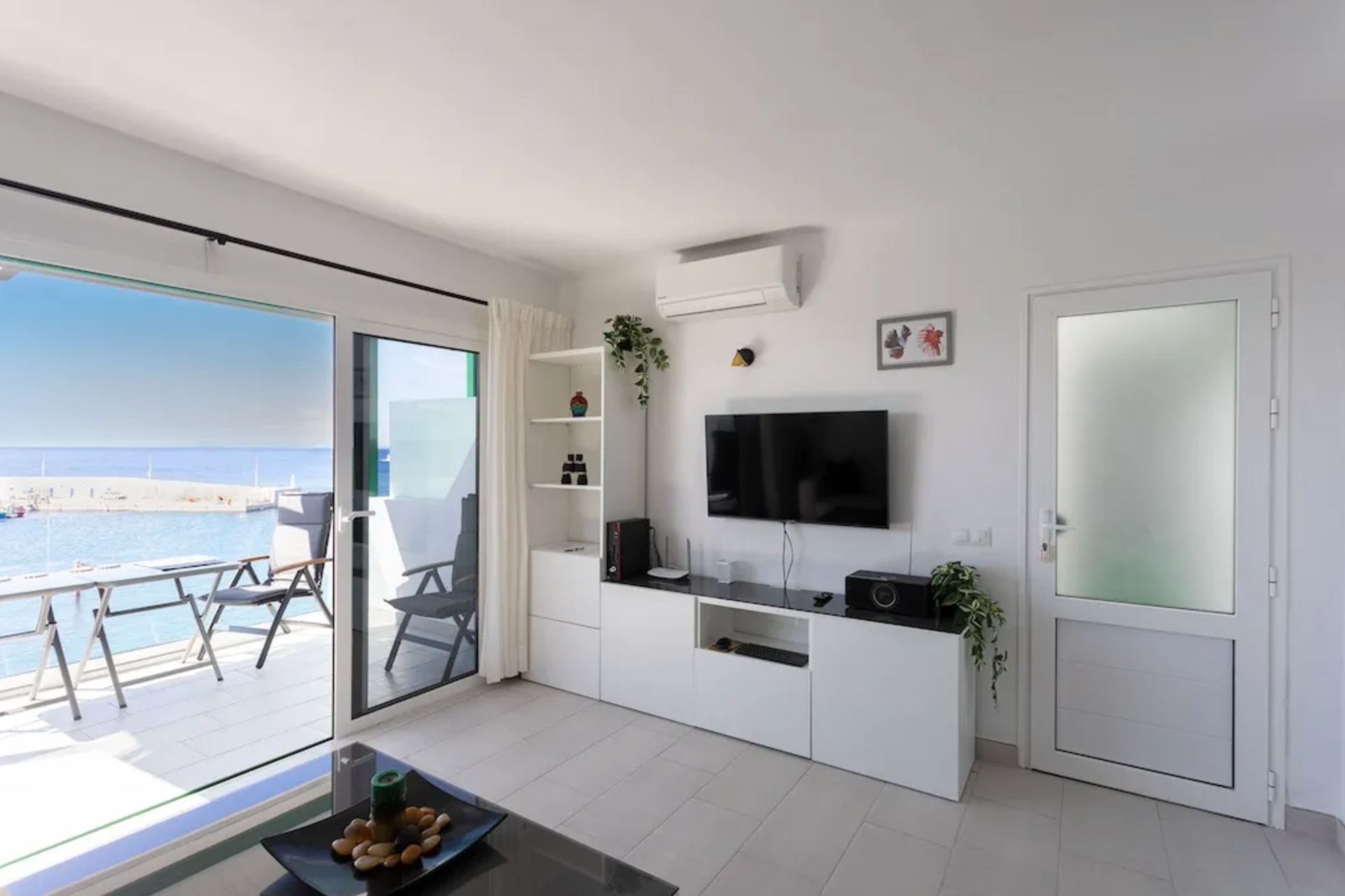 Living room with a large sliding glass door opening to a balcony overlooking the ocean, with chairs and a small table outside. Inside, there is a mounted flat-screen TV, a white entertainment unit with electronic devices, and decorative plants.