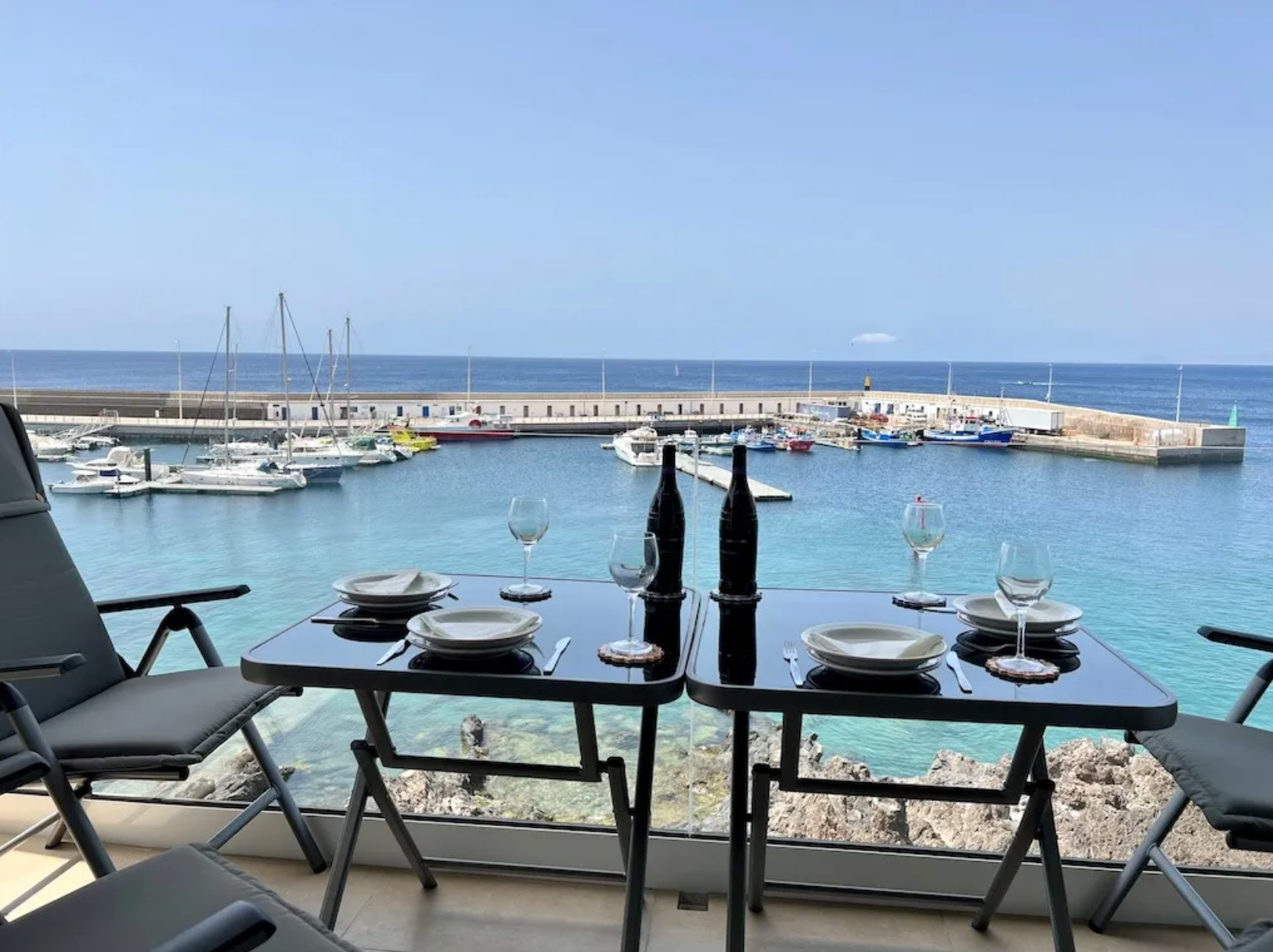 Table set for four with plates, glasses, and two bottles of wine, overlooking a marina with boats docked and an ocean view.