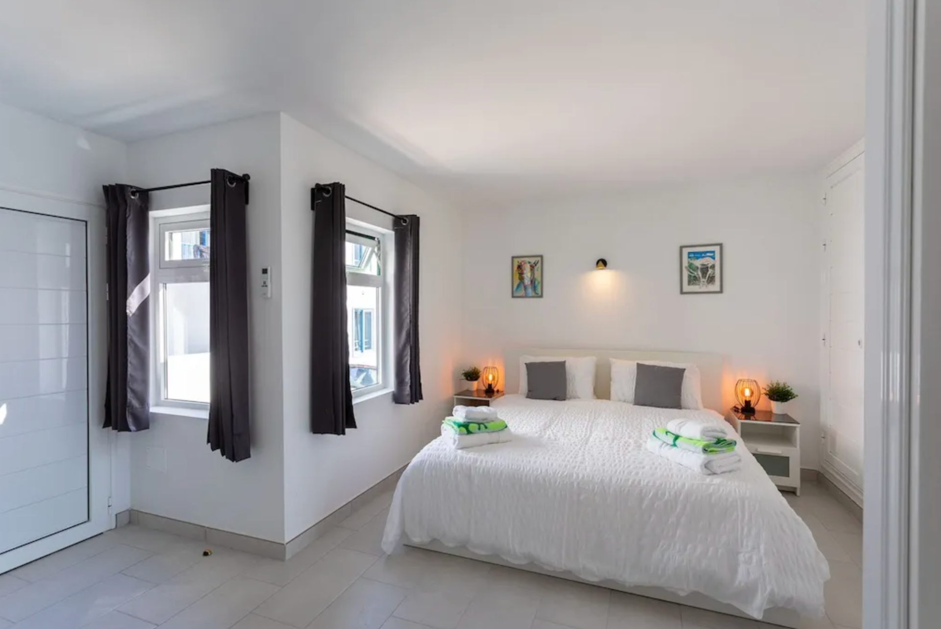 Bright bedroom with white walls, bed with white bedding and gray pillows, two nightstands with lamps, two windows with dark curtains, and two framed pictures above the bed.