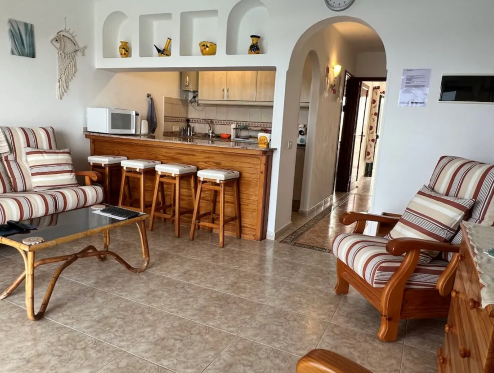 Living room with striped sofas, a glass coffee table, and a kitchen area with a bar and four barstools in a home interior.