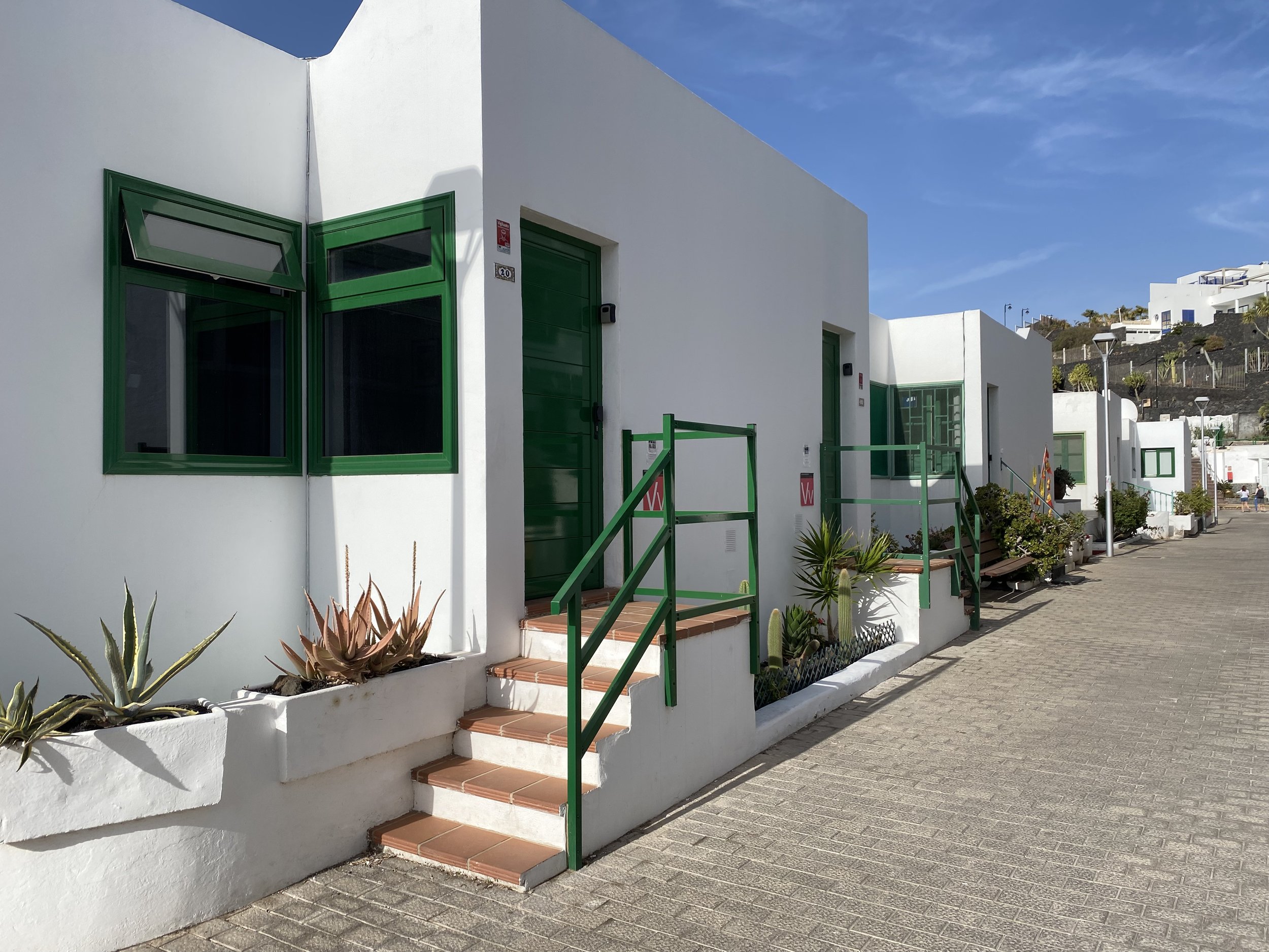 White Mediterranean-style building with green window frames, doors, and railings, along a paved walkway, with potted plants and agave in the foreground, on a sunny day with a blue sky.
