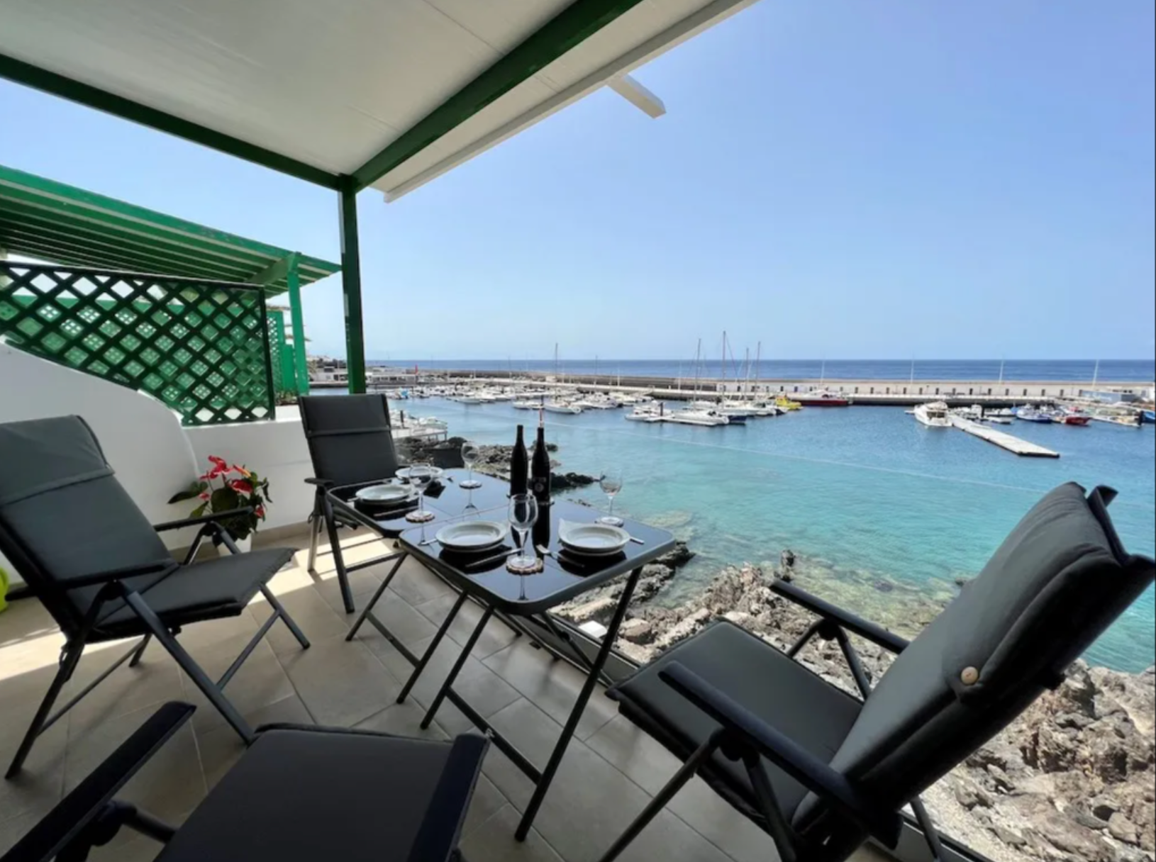 Outdoor dining area overlooking a marina with boats, table set with plates, glasses, and wine bottles, and a view of the ocean in the background.