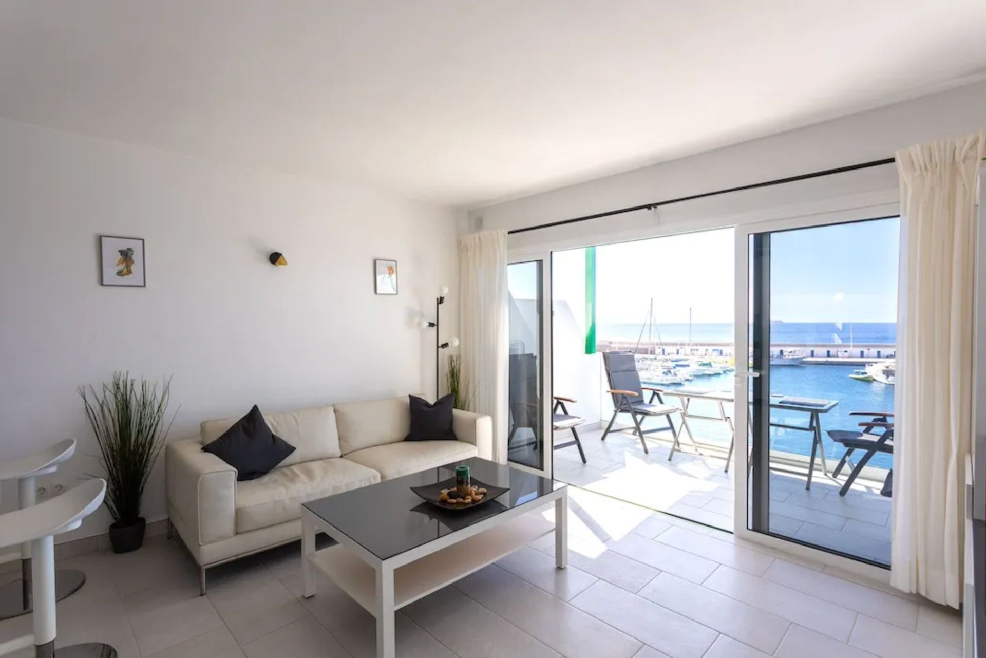 Living room with white sofa, black pillows, and sliding glass door leading to balcony with boats and ocean view.