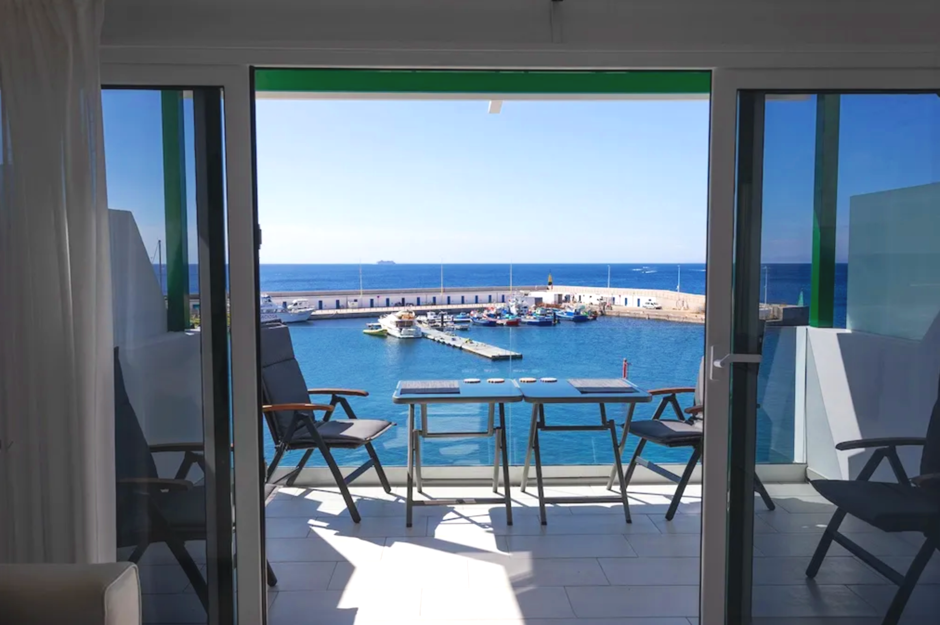 Balcony view of a marina with boats, overlooking the ocean on a sunny day, seen from inside a room with glass doors.
