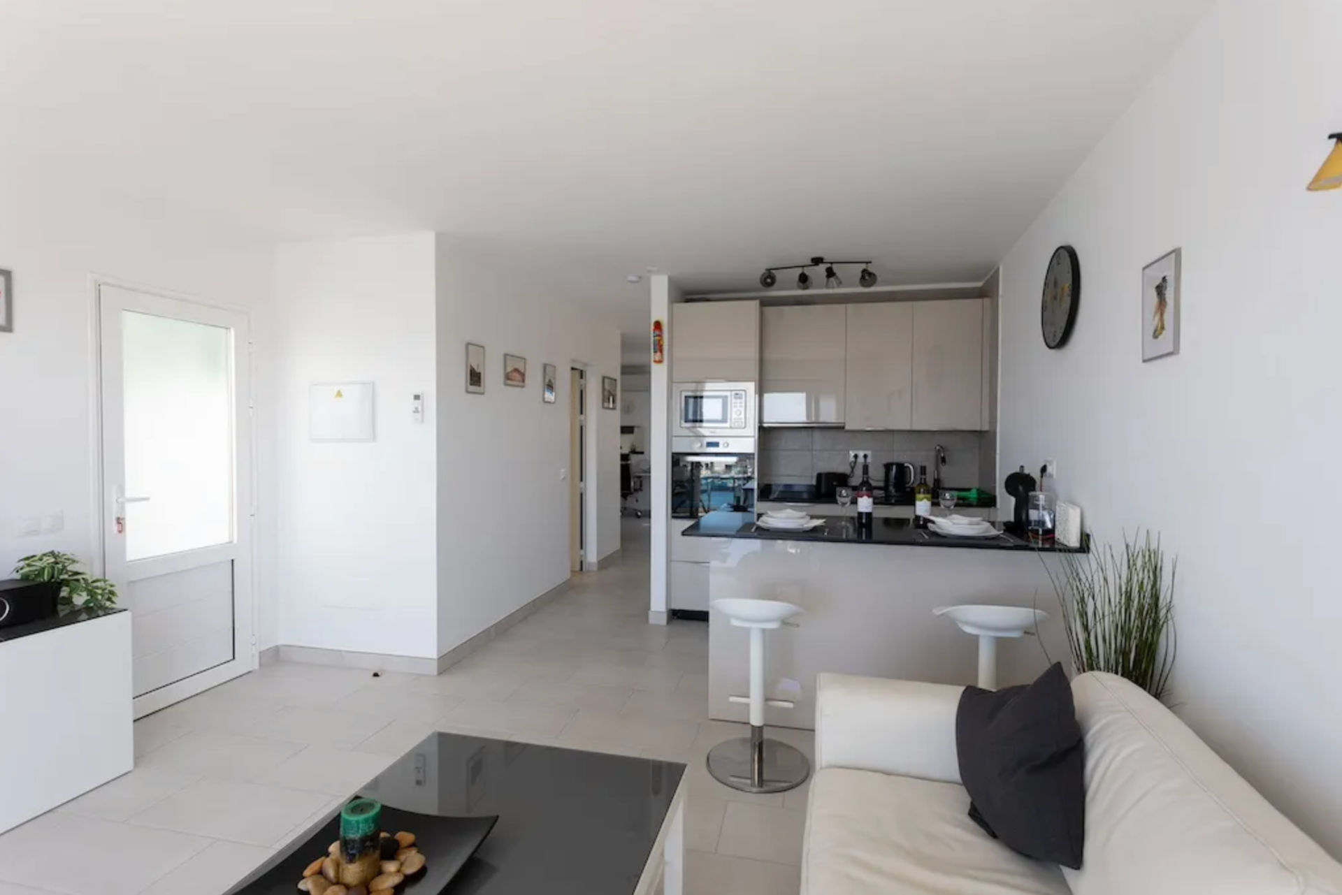 Open-plan living room with white walls and tiled floor, a beige sofa with a black cushion, a black coffee table, and a white dining counter with two white bar stools. The kitchen area features beige cabinets, a microwave, and various small appliances