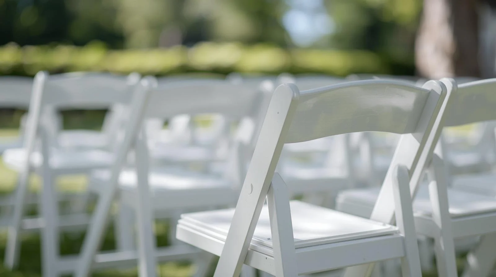 Rows of white folding chairs set up outdoors for an event, with blurred green trees and grass in the background.