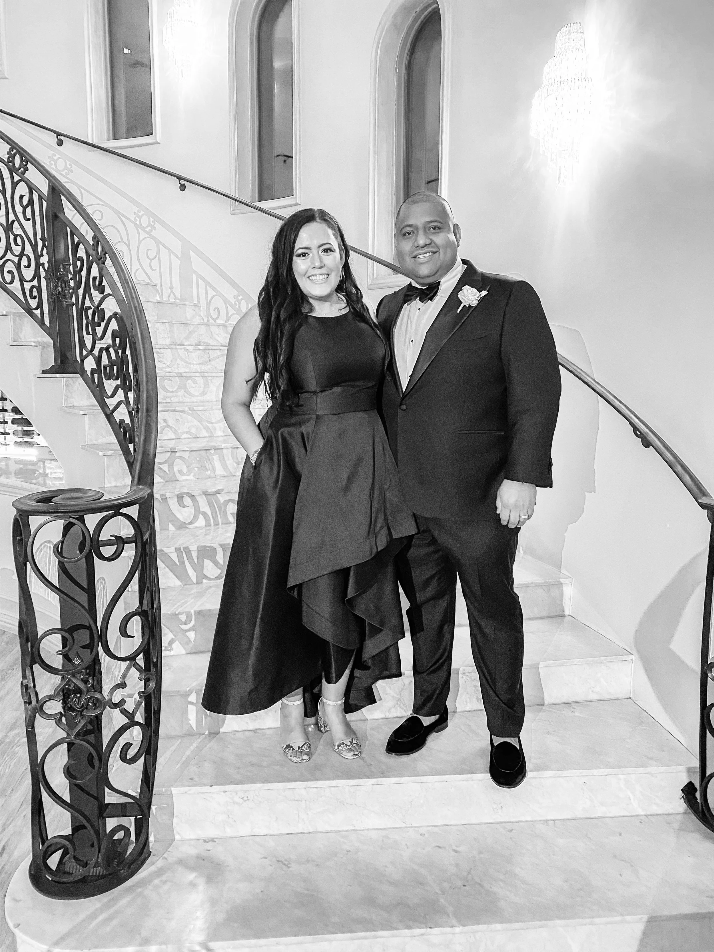 A man and woman dressed in formal attire standing on a staircase, smiling for the photo, with elegant iron railings and a chandelier in the background.