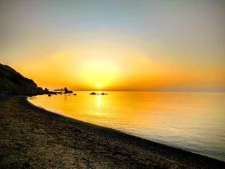 Sunset over a calm beach with a rocky shoreline on the left and smooth water on the right. From Dr. Michael Kellis's village in Greece.