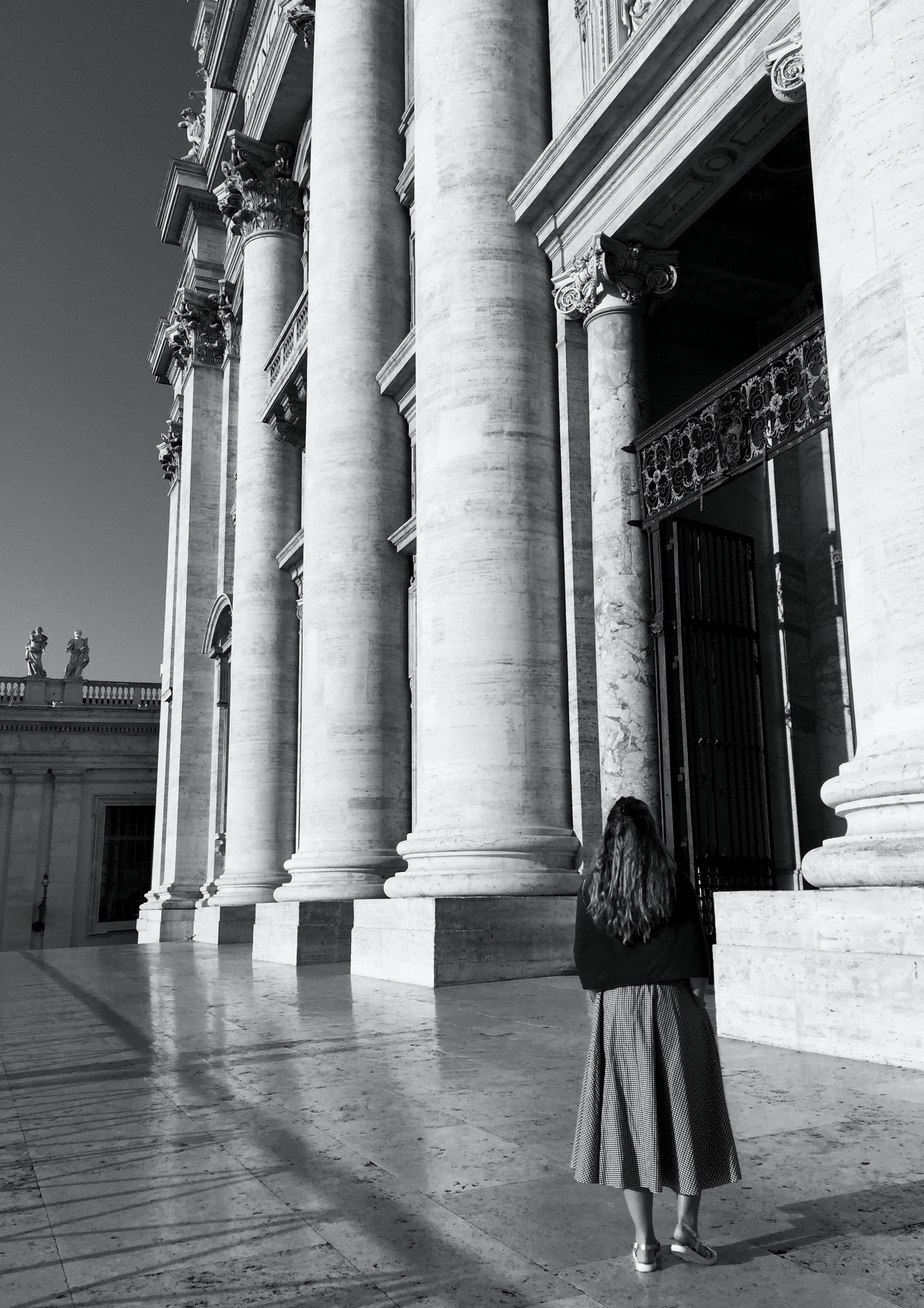 Une femme regarde vers une grande façade de bâtiment avec de grosses colonnes en pierre, dans un style architectural classique, en noir et blanc.