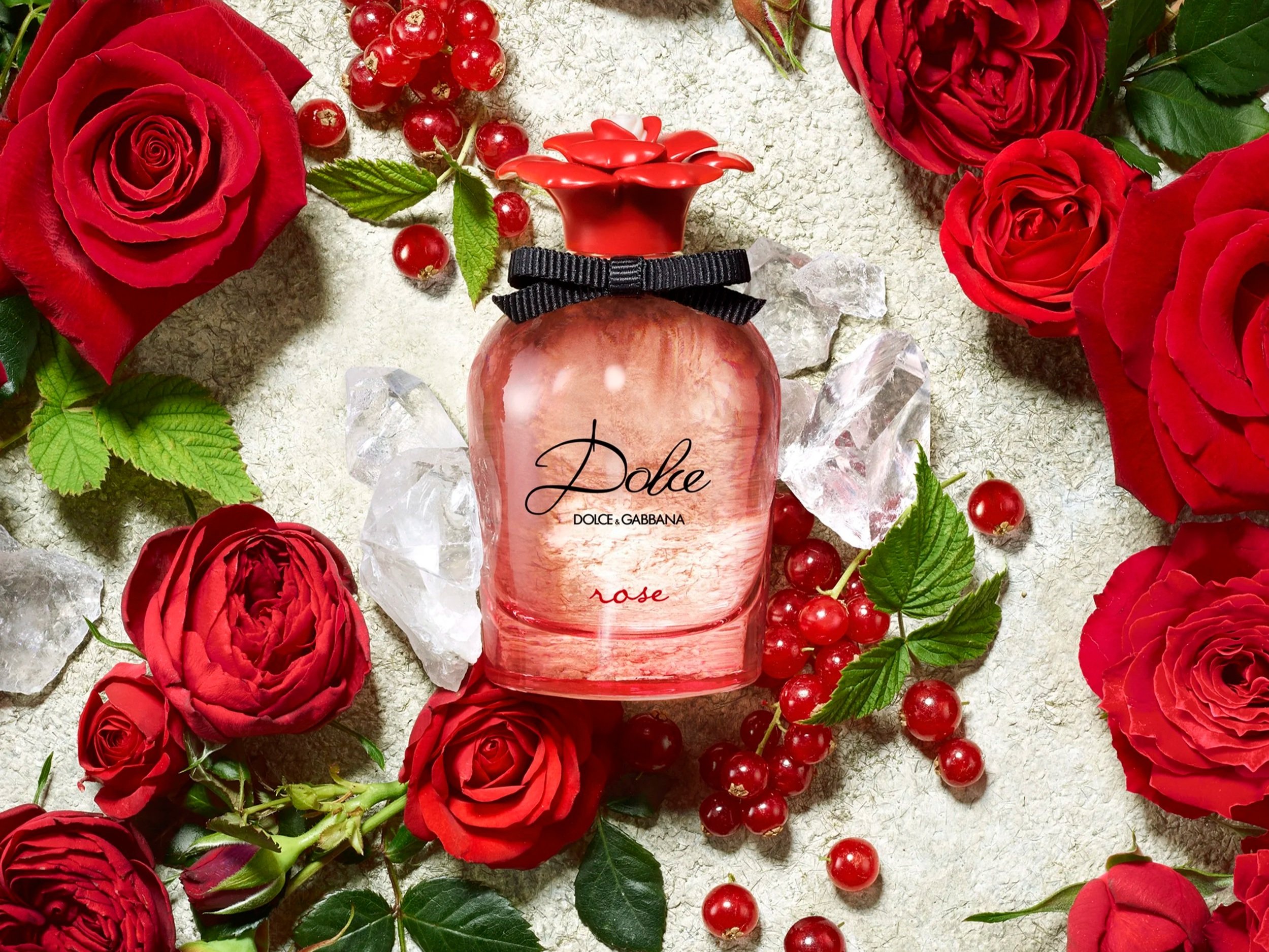 Une bouteille de parfum rose au centre, entourée de roses rouges, de groseilles rouges, et de morceaux de glace, sur une surface beige.