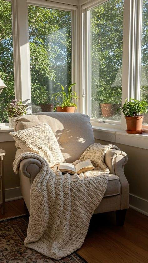 Sunlit cozy corner of a living room featuring a beige armchair with a knit blanket and pillow, an open book, and four potted plants on the window sill, with large windows showing green trees outside.