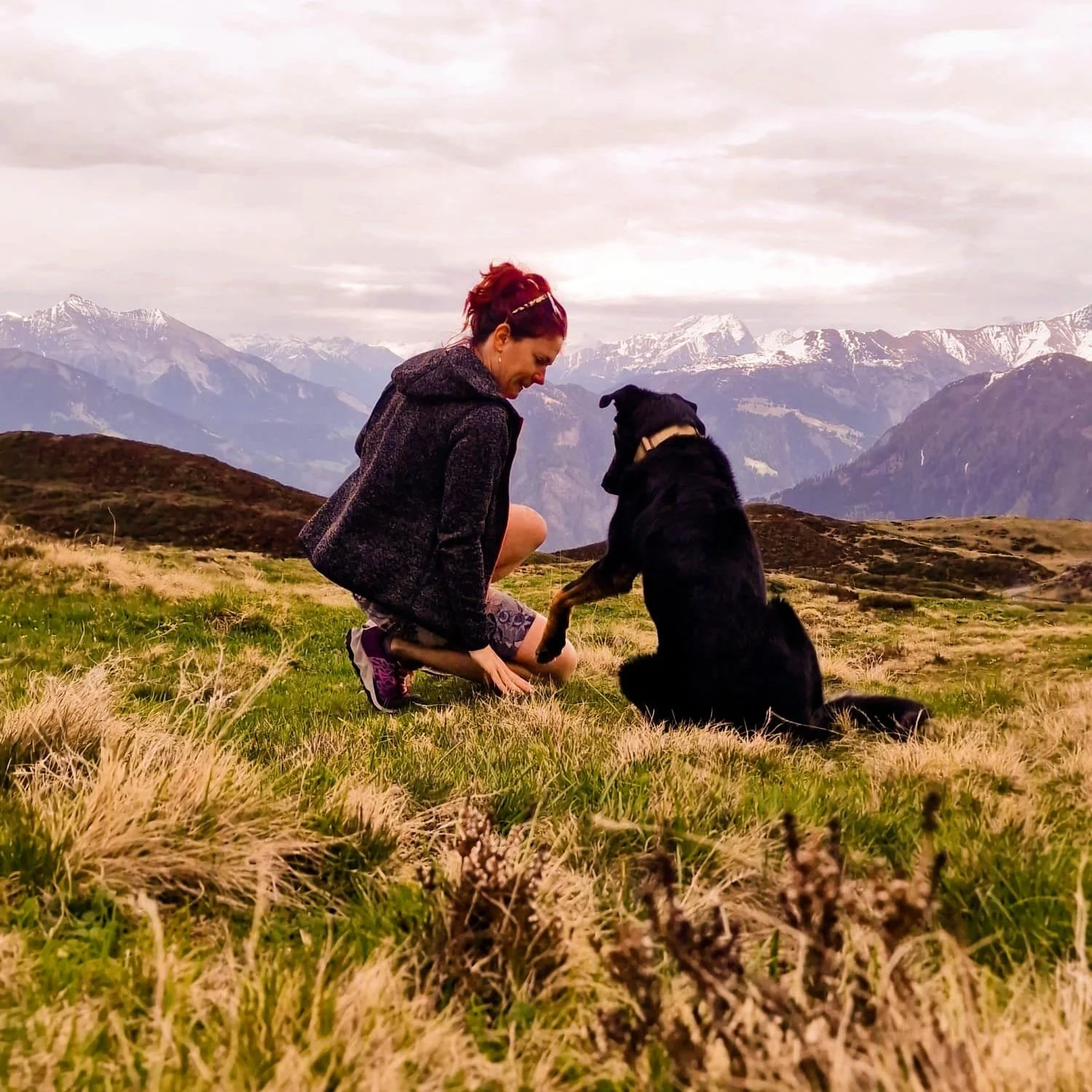 Melanie Safret und Miri auf einer grünen Wiese in den Bergen, umgeben von Schnee bedeckten Gipfeln. Die Frau kniet und spricht zu dem sitzenden Beauceron.