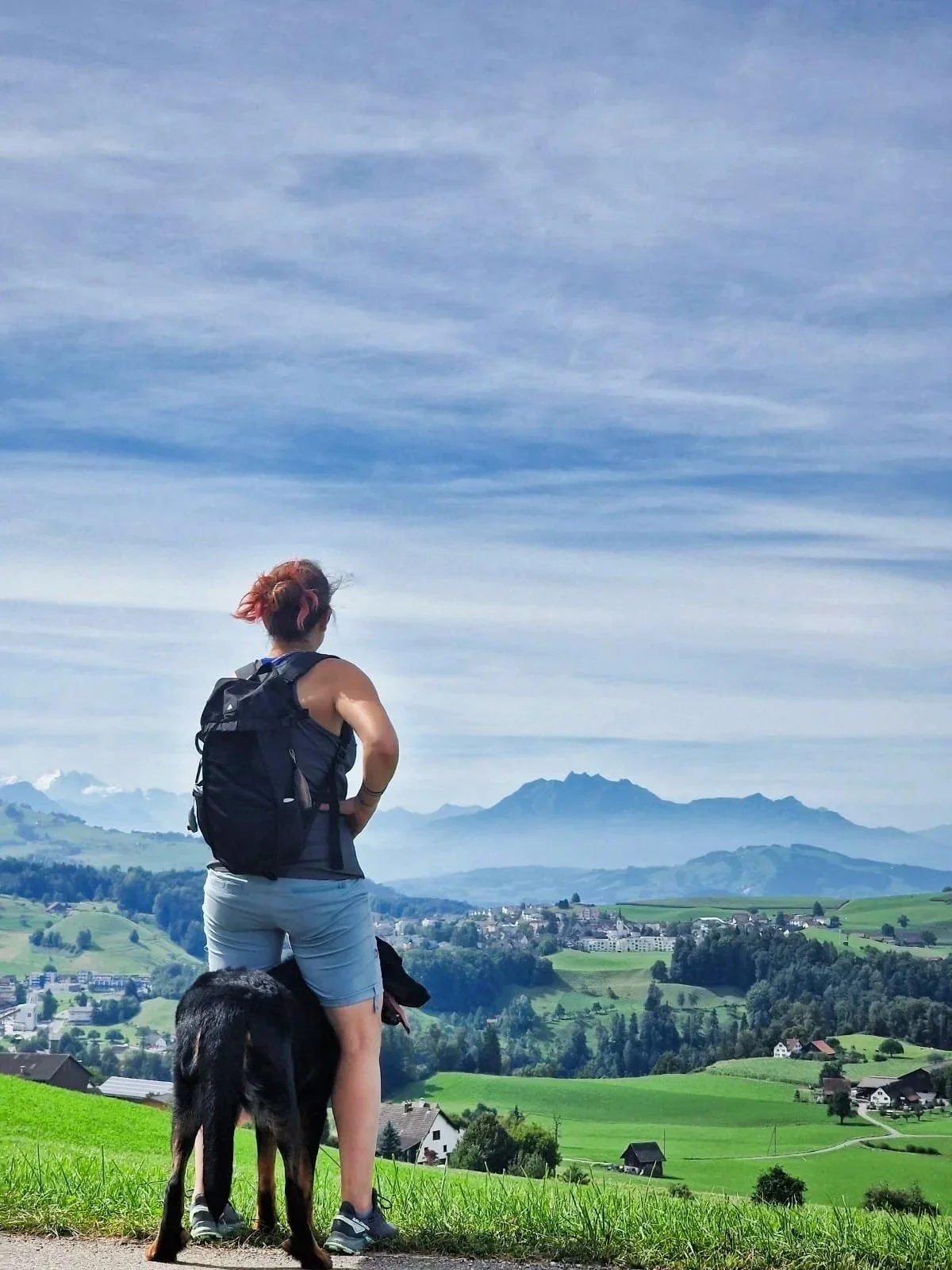 Melanie mit Rucksack steht mit einem schwarzen Hund auf einer grünen Wiese und betrachtet eine Berglandschaft mit Häusern und Hügeln unter einem blauen Himmel.