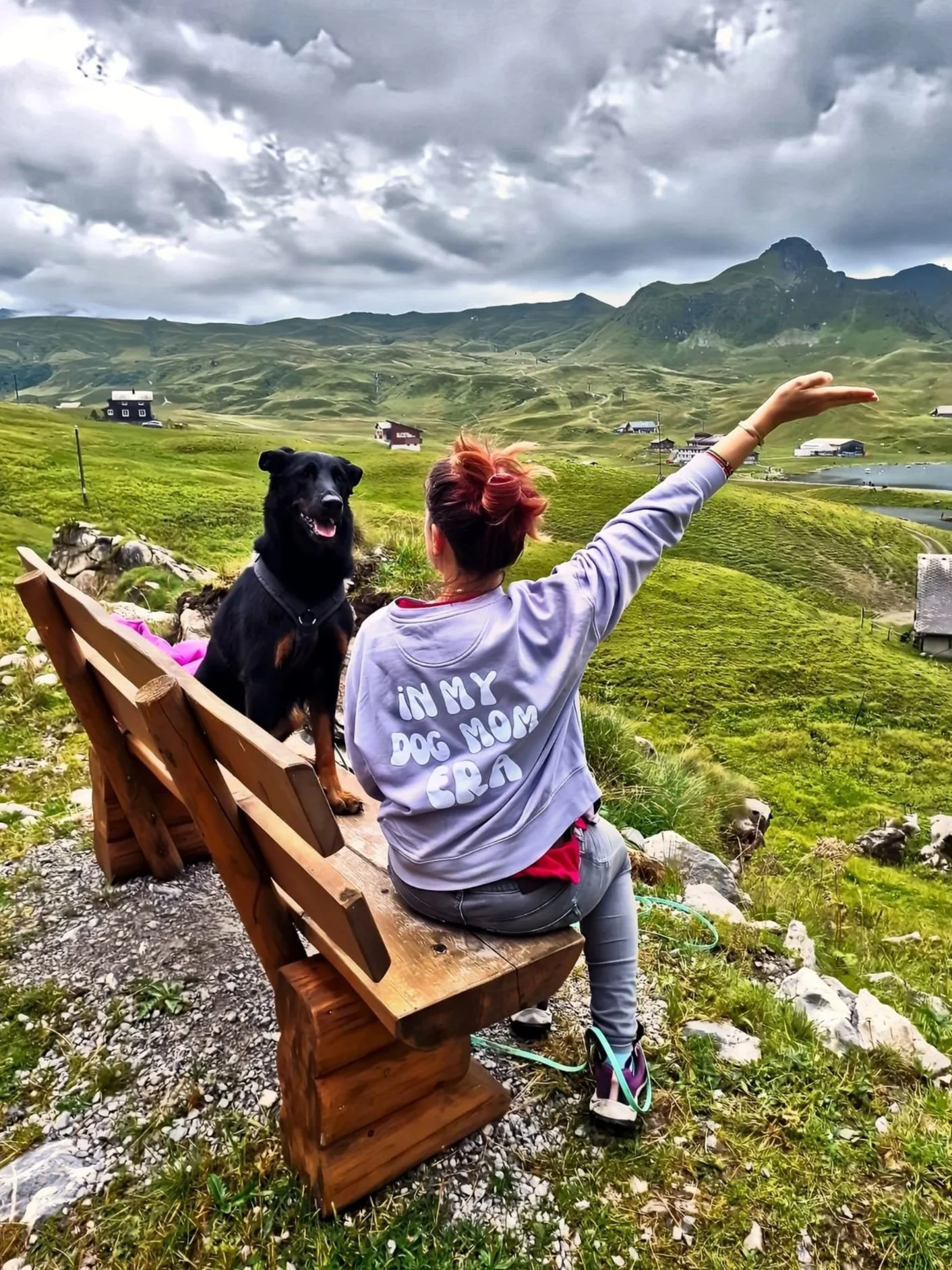 Mel sitzt auf einer Holzbank und schaut auf eine Berglandschaft mit Hütten, Wasser und bewölktem Himmel. Neben ihr sitzt Miri,  Beauceron.