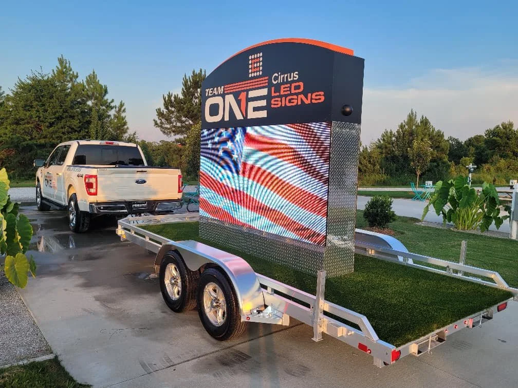 A white pickup truck parked next to a trailer with a large digital LED sign displaying patriotic red, white, and blue wave patterns, with trees and a blue sky in the background.