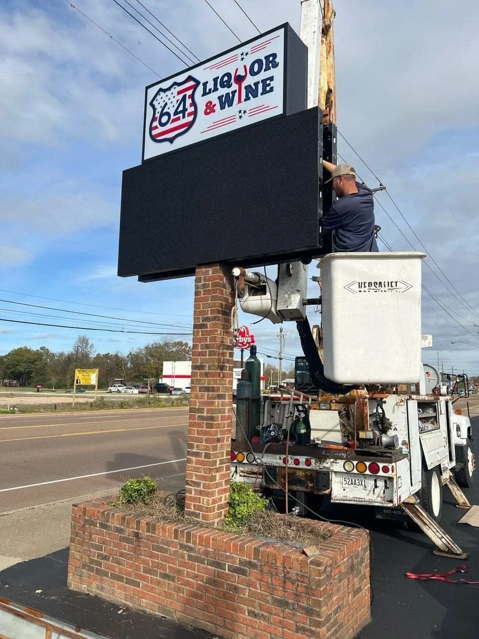 A worker on a lift station installing or repairing a large digital sign for '64 Liquor & Wine' on the side of a brick planter along a roadside.