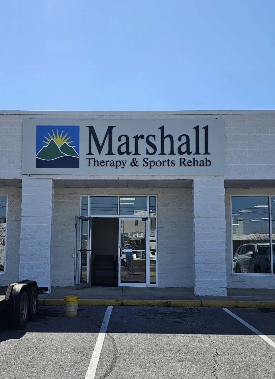 The storefront of Marshall Therapy & Sports Rehab with a sign featuring mountains and a sun, two parking spaces in front, and a clear blue sky above.