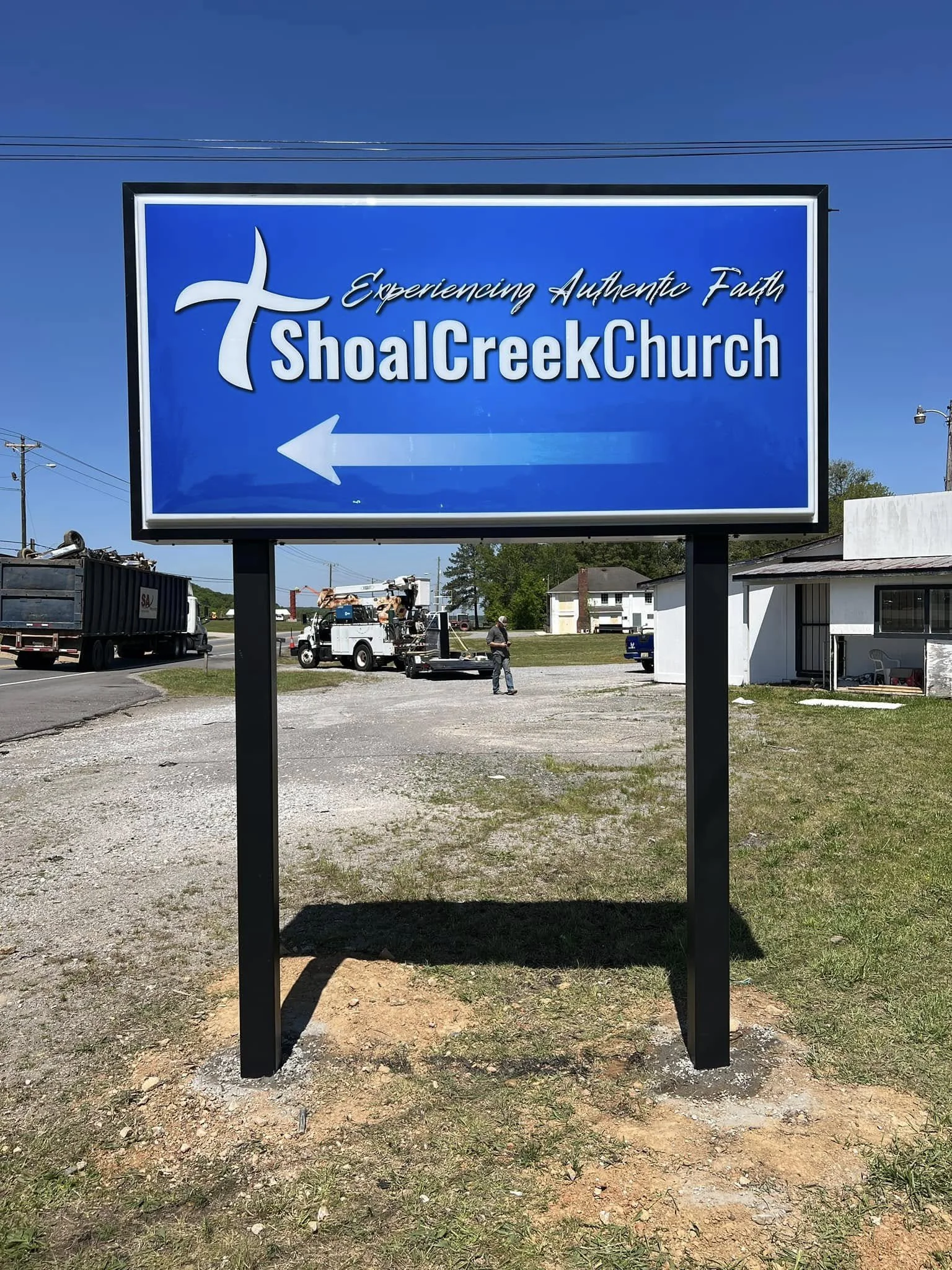 A large blue sign for Shoal Creek Church with white text and a white cross symbol, featuring an arrow pointing left.