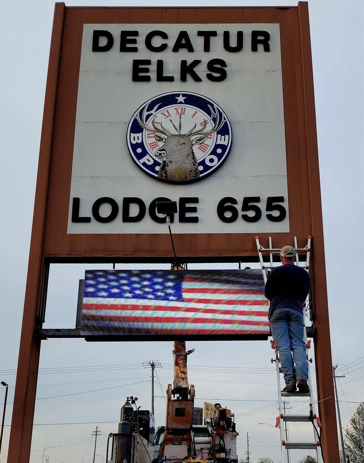 A worker on a ladder installing or repairing a digital display on a large sign for Decatur Elks Lodge 655, with an American flag graphic on the screen.