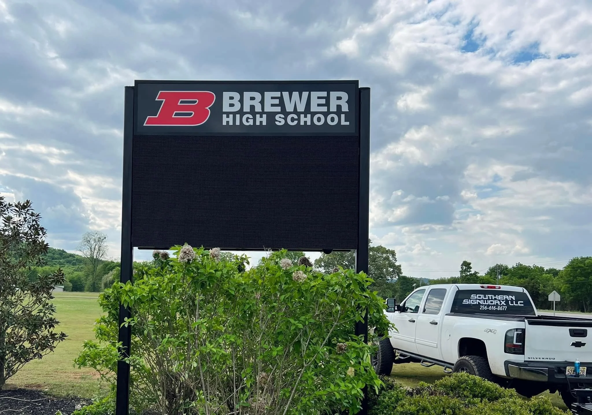 A school sign for Brewer High School with a black electronic display board below the school name, surrounded by green bushes. A white pickup truck is parked nearby, with the company's name 'Southern Sign Worx LLC' on the back window. The sky is partl