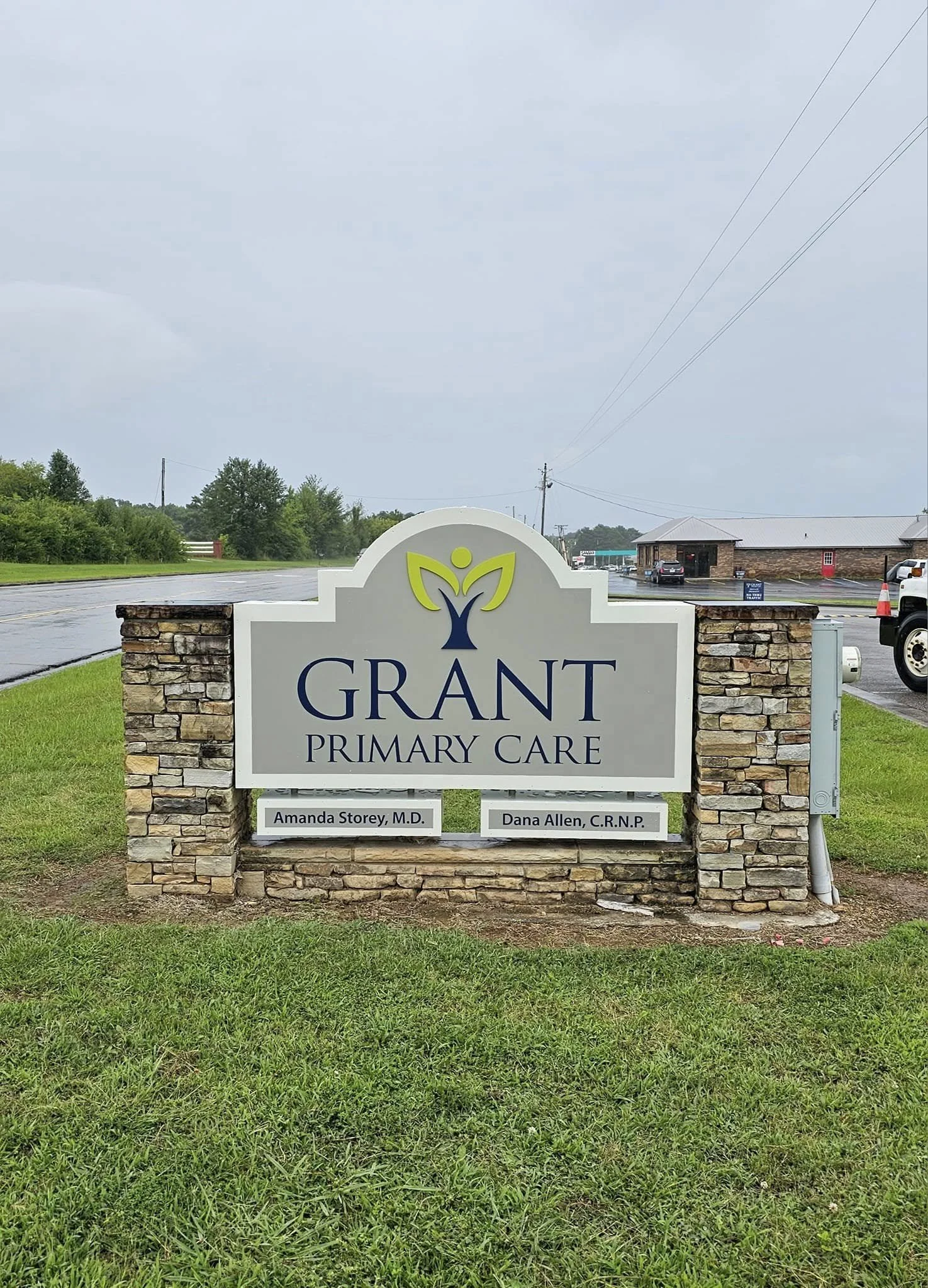 Sign for Grant Primary Care with names Amanda Storey, M.D., and Dana Allen, C.R.N.P. in front of a brick and stone structure, on a grassy area near a road under a cloudy sky.