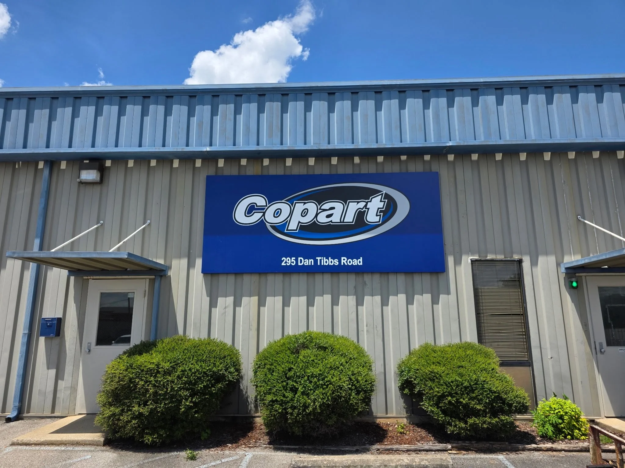 Exterior of a Copart auto auction facility with a blue sign on a silver metal building, three green bushes in front, a small door to the left, and a window to the right. The sky is blue with some clouds.