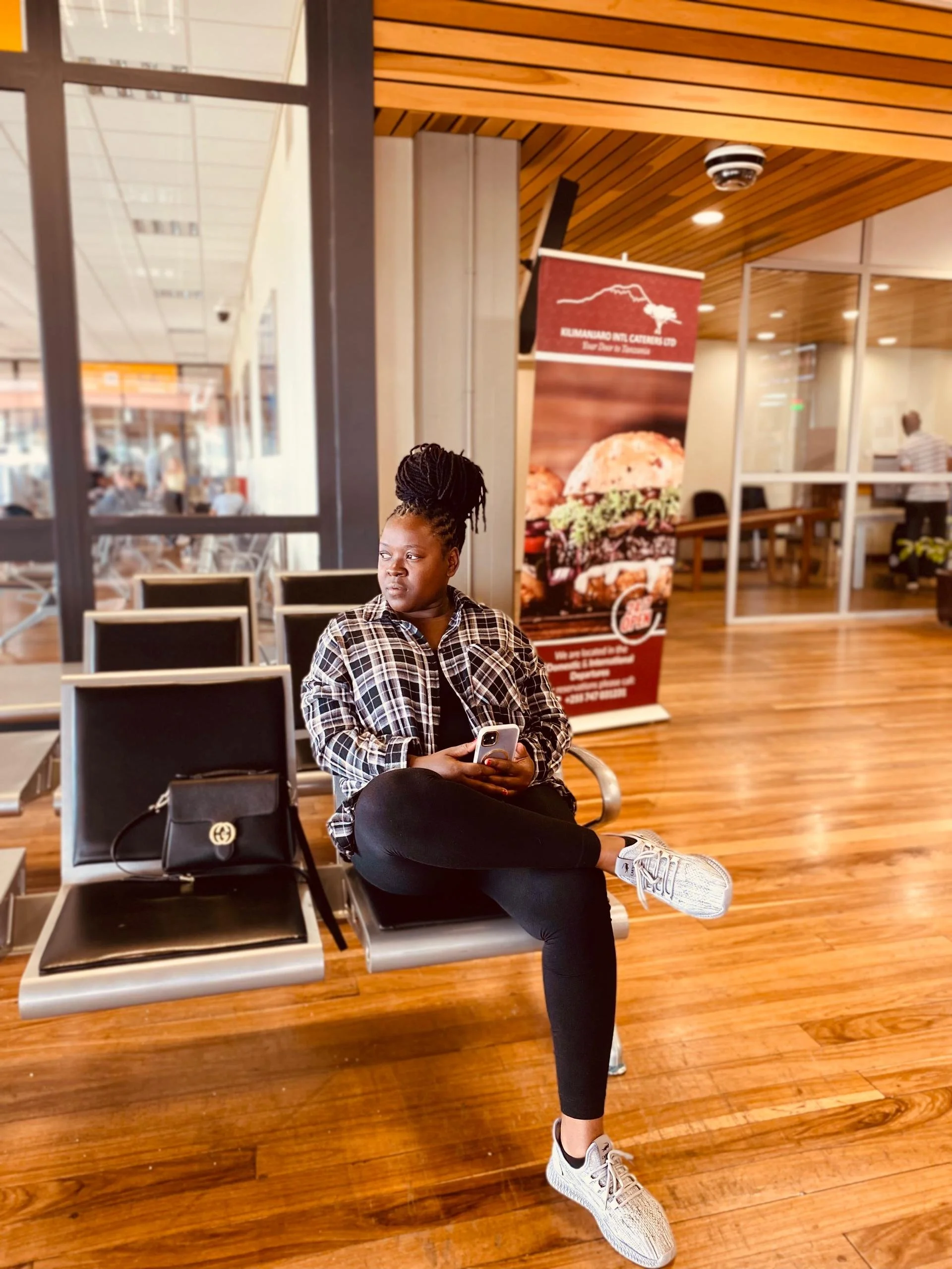 A woman with braided hair tied in a bun, sitting on a black bench at an airport terminal, looking to her left and holding a smartphone. She is wearing a checkered shirt, black leggings, and white athletic shoes. There is a black handbag placed on the bench beside her. In the background, there is a poster advertising a restaurant and a glass wall with people walking outside.