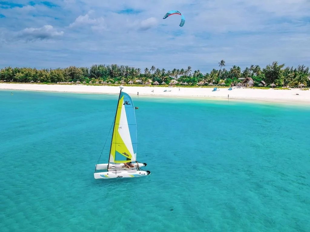 A sailboat with a yellow and white sail on turquoise water near a white sandy beach with palm trees and thatched-roof huts.