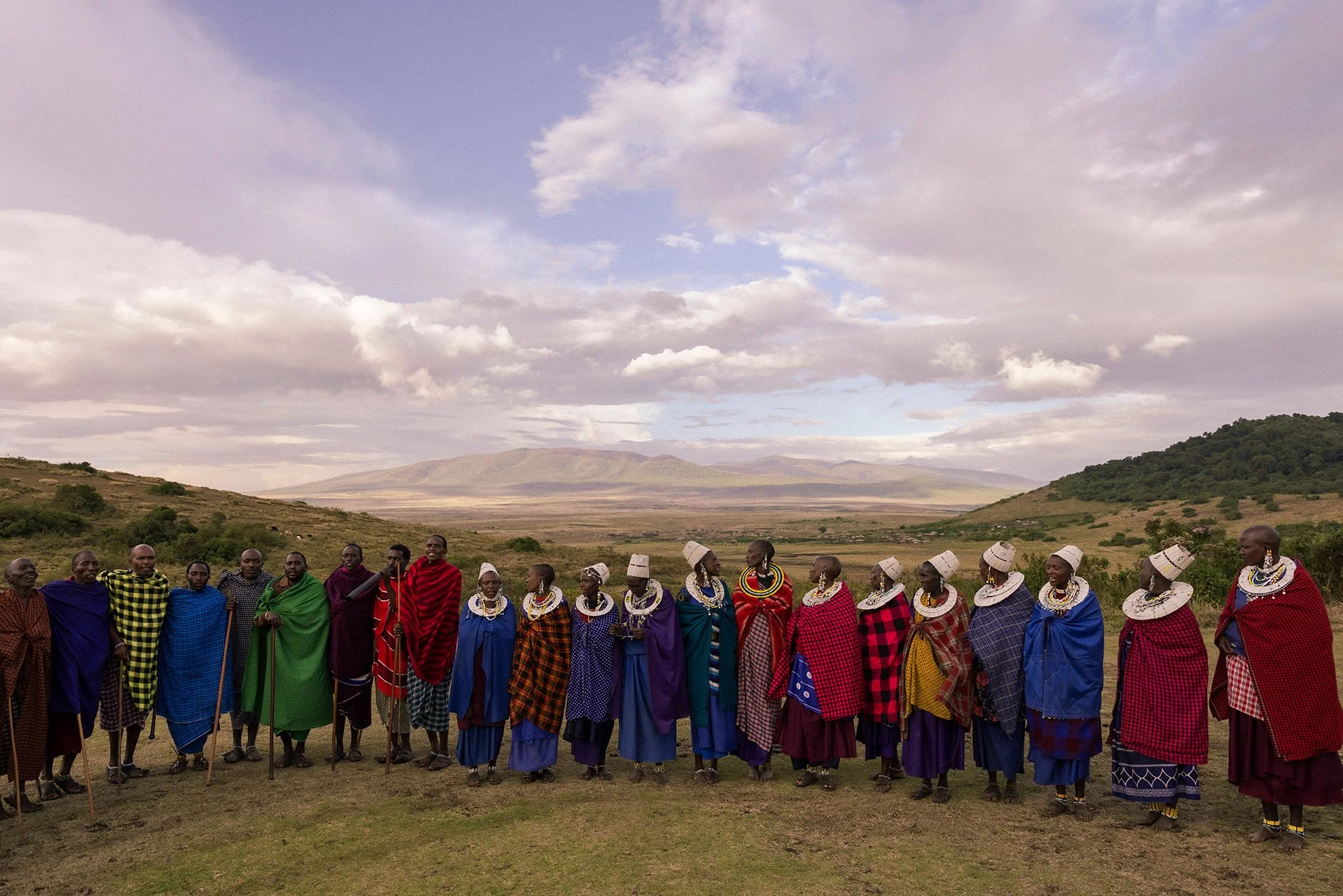 Group of Maasai people standing outdoors in a line, wearing traditional colorful clothing and jewelry, with a vast landscape and mountains in the background.